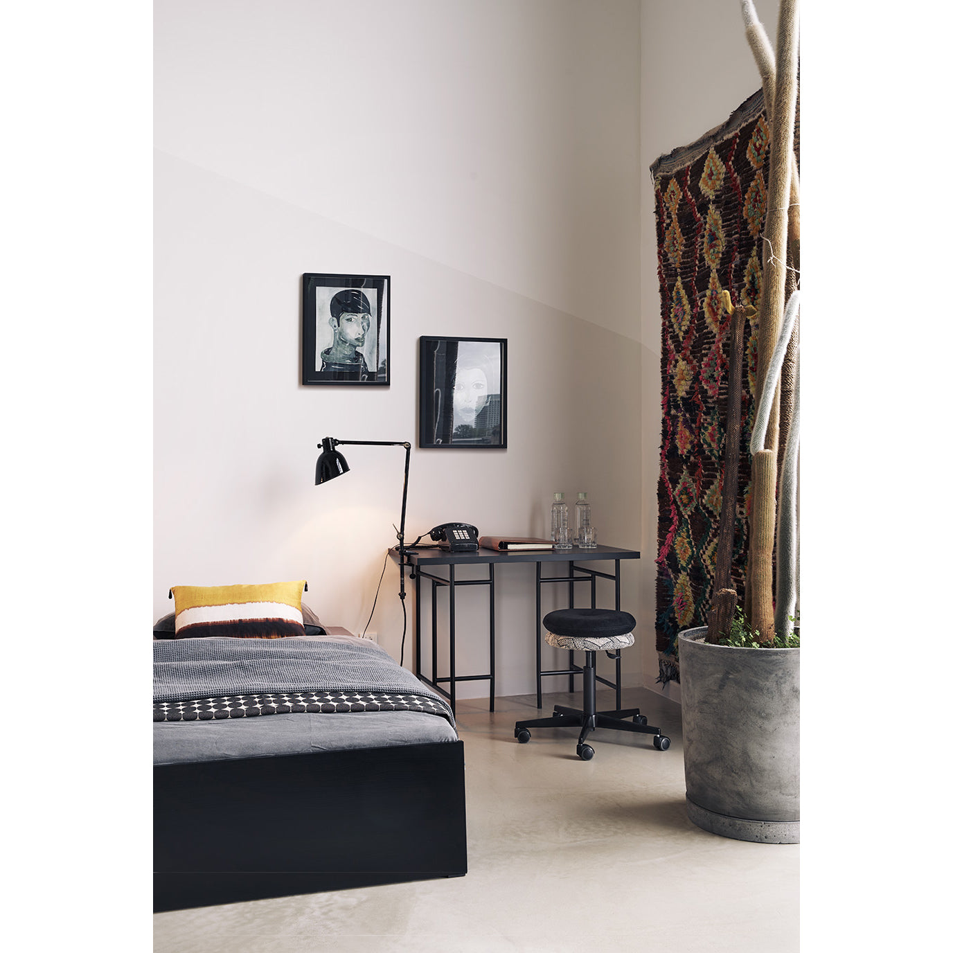A minimalist bedroom features a single bed with gray bedding and a yellow pillow. Nearby is a black desk with a lamp, chair, and the Mr. Round Swivel Stool from Richard Lampert, featuring a gas spring system for height adjustment. Wall art and potted plant by multicolored curtains complete the look.
