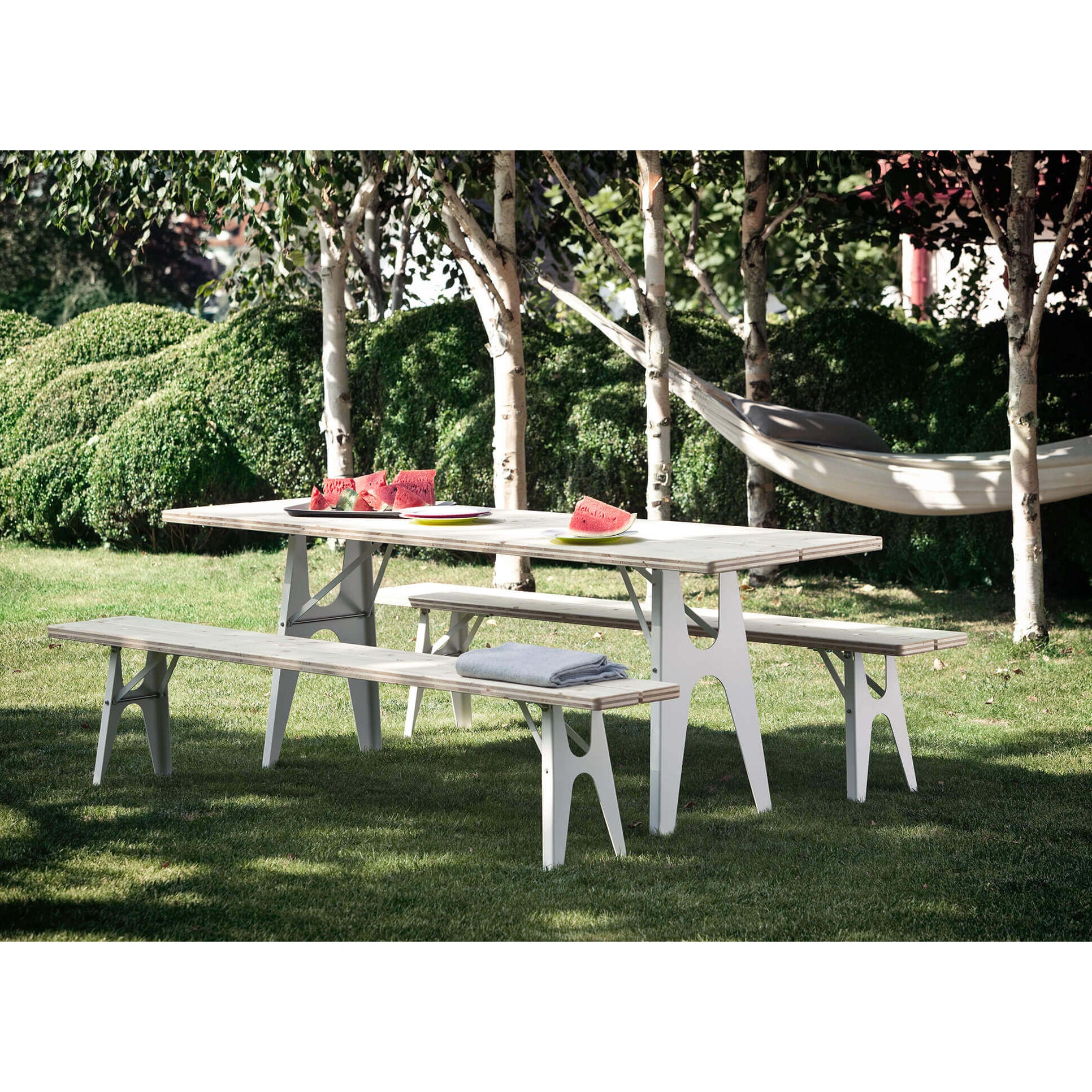 The Ludwig Bench by Richard Lampert, crafted for outdoor use, sits on a grassy lawn encircled by trees. Watermelon slices are on the bench alongside a folded blanket. A hammock sways between two trees in the background, creating an ideal setting of relaxation and nature.