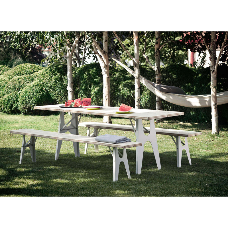 The Ludwig Bench by Richard Lampert, crafted for outdoor use, sits on a grassy lawn encircled by trees. Watermelon slices are on the bench alongside a folded blanket. A hammock sways between two trees in the background, creating an ideal setting of relaxation and nature.