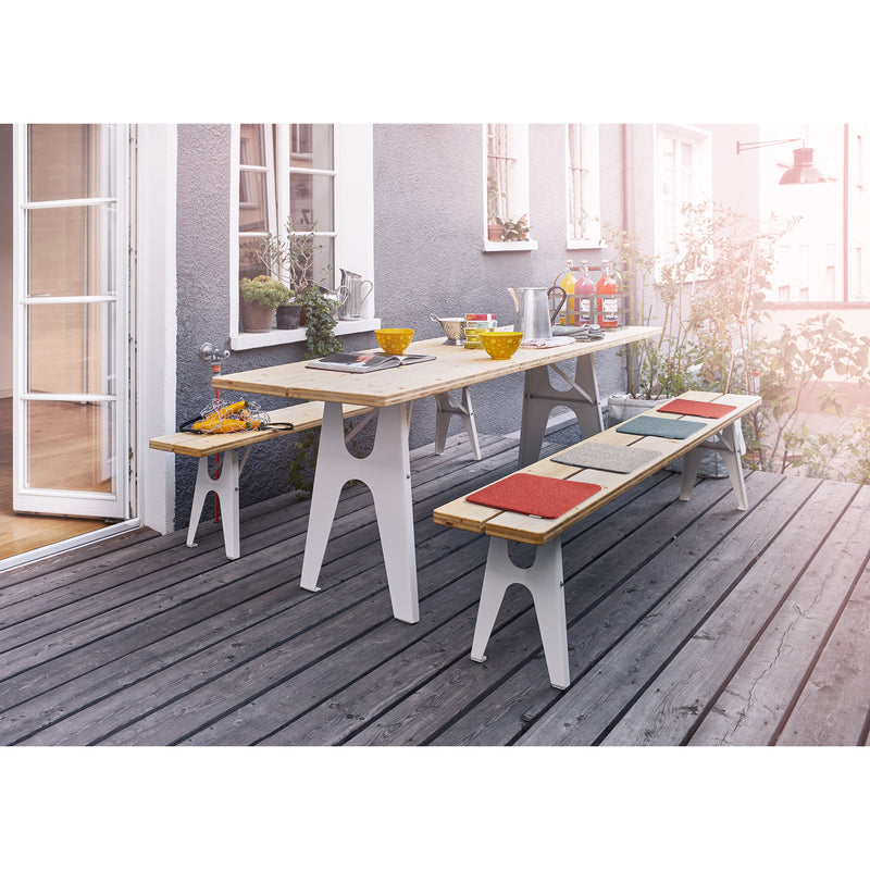 The outdoor dining area features a sustainable design with a wooden table and Richard Lamperts Ludwig Benches on the deck. This inviting beer table is set with yellow bowls, drinks, and tableware, while the colorful-cushioned benches are surrounded by lush plants next to a windowed door.