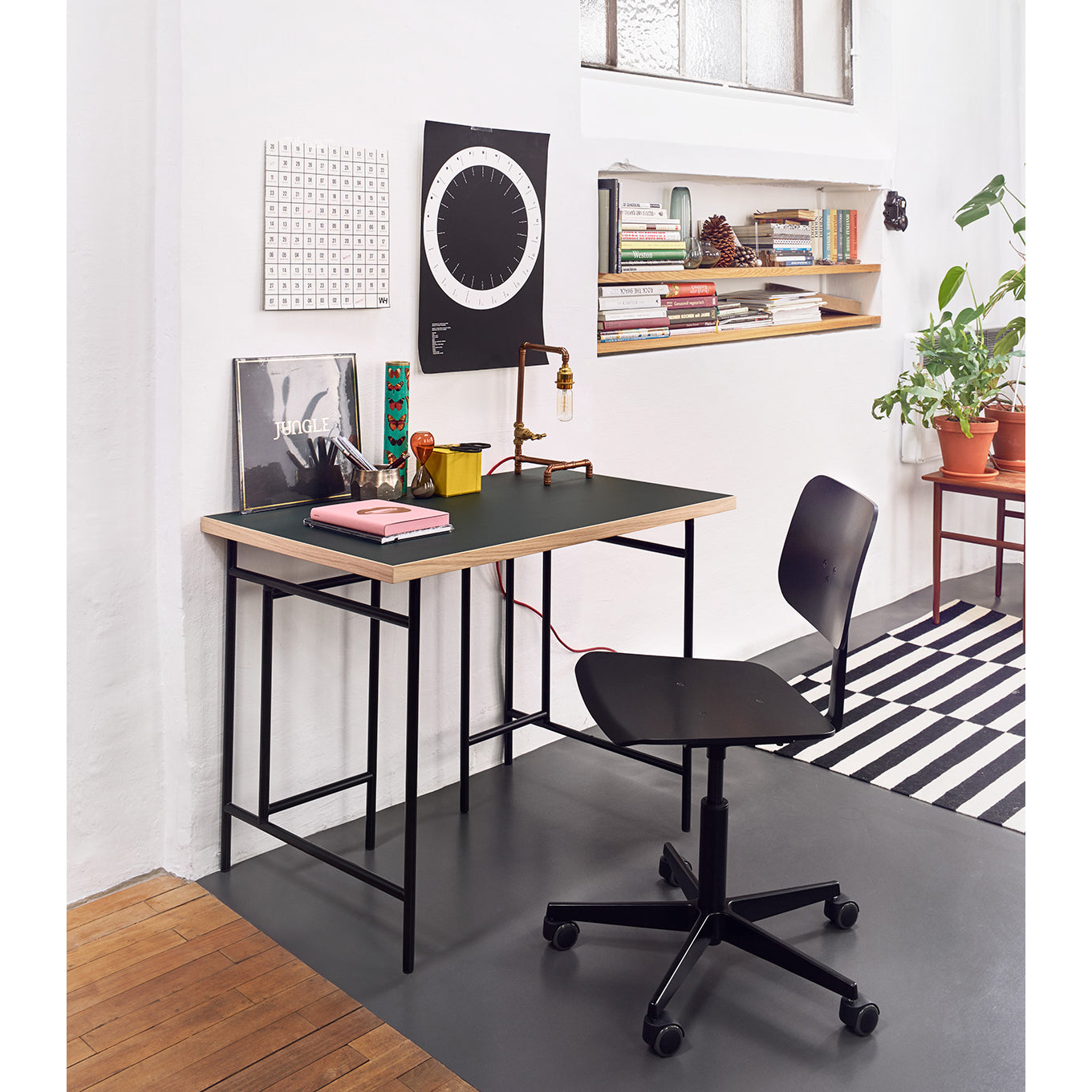A compact home office setup includes a sleek desk paired with the Mr. Square Task Chair by Richard Lampert, offering superior comfort. The desk holds a lamp, books, and decor while a black and white wall clock, art, and small potted plants add a modern touch atop the wooden floor with striped rug accents.