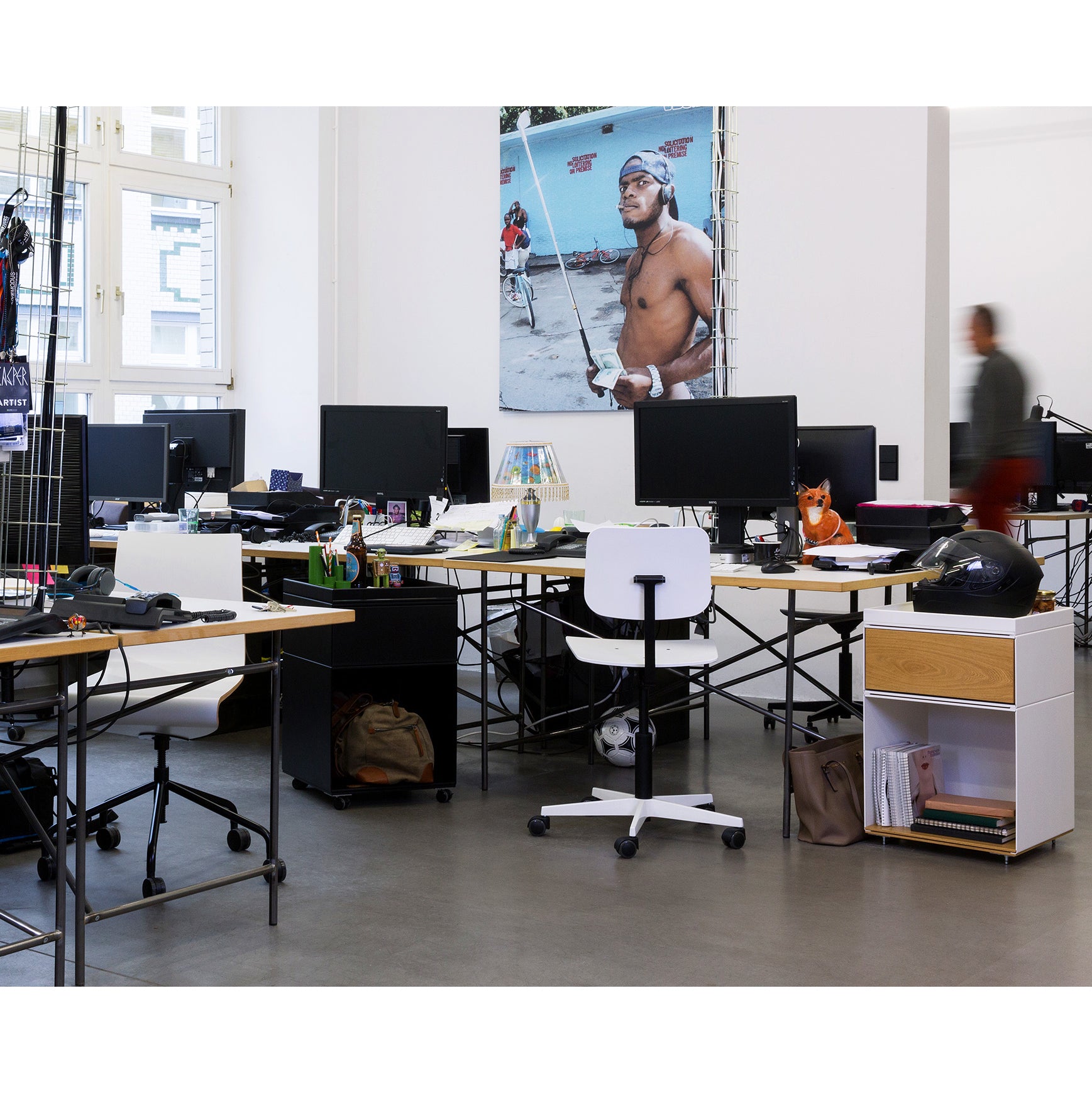 A modern office features multiple desks with computers and Richard Lamperts Mr. Square Task Chairs for superior comfort, along with a poster of a muscular person on a beach. A blurred figure walks in the background, and natural light floods through large windows.
