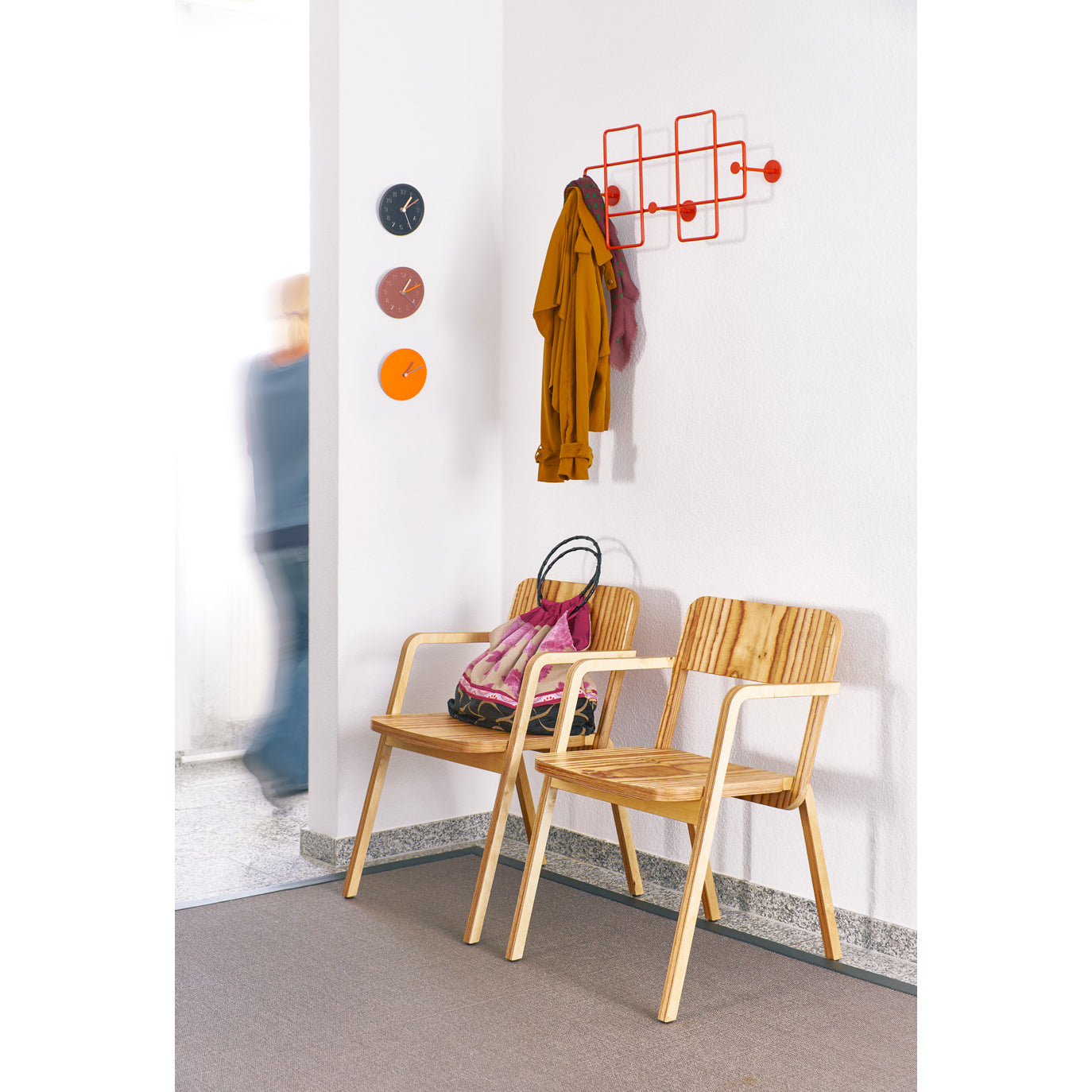 A minimalist waiting area includes two wooden chairs with a bag on one, colorful clocks, and a scarf near sleek Bazar Coat Racks by Richard Lampert. A blurred person injects dynamic energy into the modern home organization space.