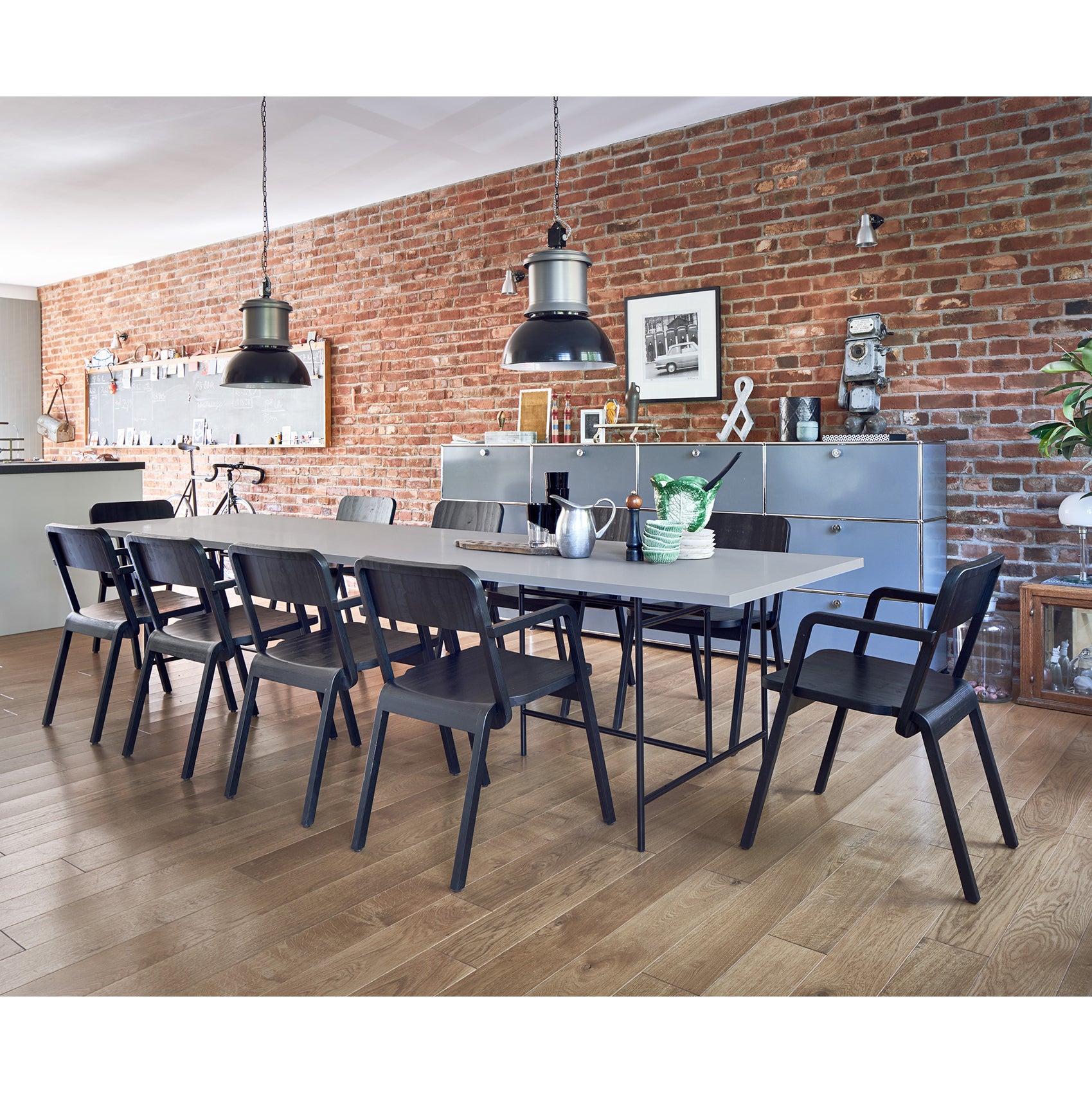 In a modern dining area, a long table is surrounded by ten Prater Chairs from Richard Lampert on a wooden floor. A brick wall highlights two large hanging lamps, metallic cabinets, and decorative items. Natural light enhances the inviting atmosphere.