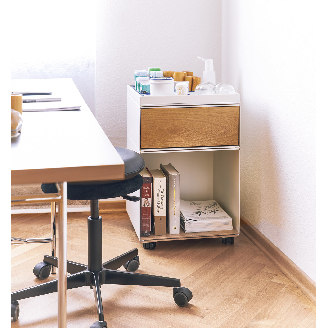 The Stak Trolley System by Richard Lampert, a compact white rolling cart with adjustable castors and a wooden drawer, sits in the room corner. Its top holds containers and sanitizer; below, books and a magazine are neatly arranged. Nearby, part of a table and chair are visible.