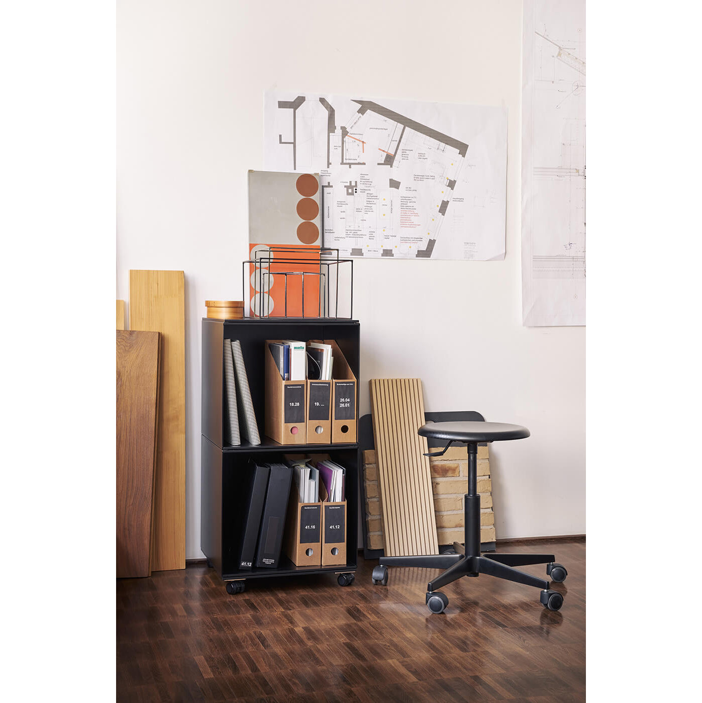 In an office corner, the Stak Trolley System by Richard Lampert organizes files and notebooks with its black shelving. Architectural drawings are pinned above. Solid oak shelves store wood planks and tiles adjacent to it. A black swivel stool sits on the wooden floor.