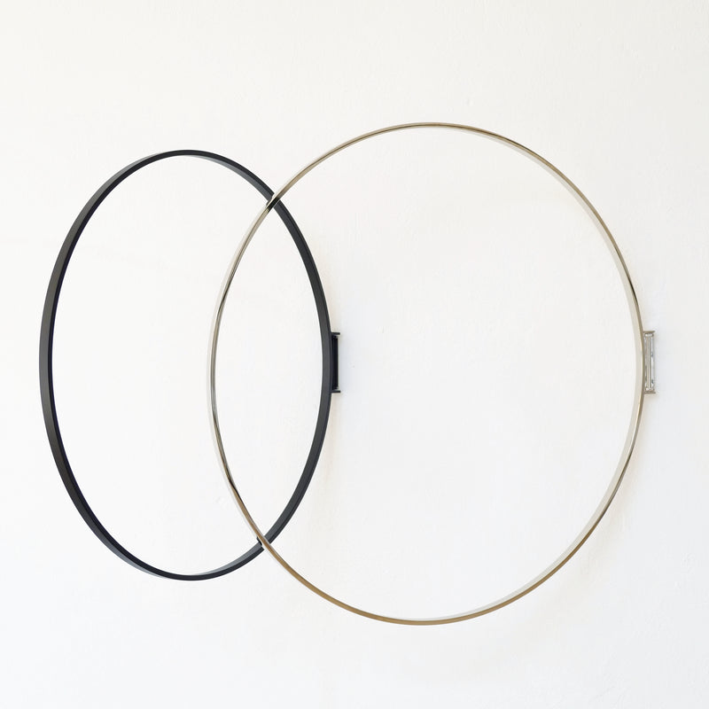Two large metal rings, one black and one silver, are mounted on a white wall by Atelier Haussmanns Rings, slightly intersecting. A modern geometric design with shadows emerges, creating a stylish coatrack that functions as both decor and utility.