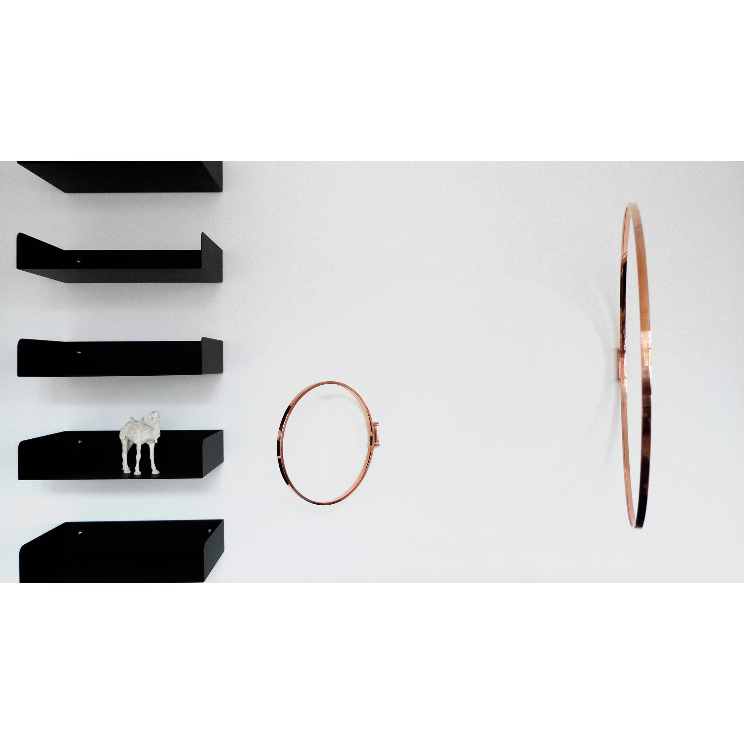 A minimalist interior showcases vertical black shelves against a white wall. A small animal figurine sits on the second shelf from the top, while two metal Rings by Atelier Haussmann, one larger and one smaller, are elegantly mounted as decorative objects on the wall.