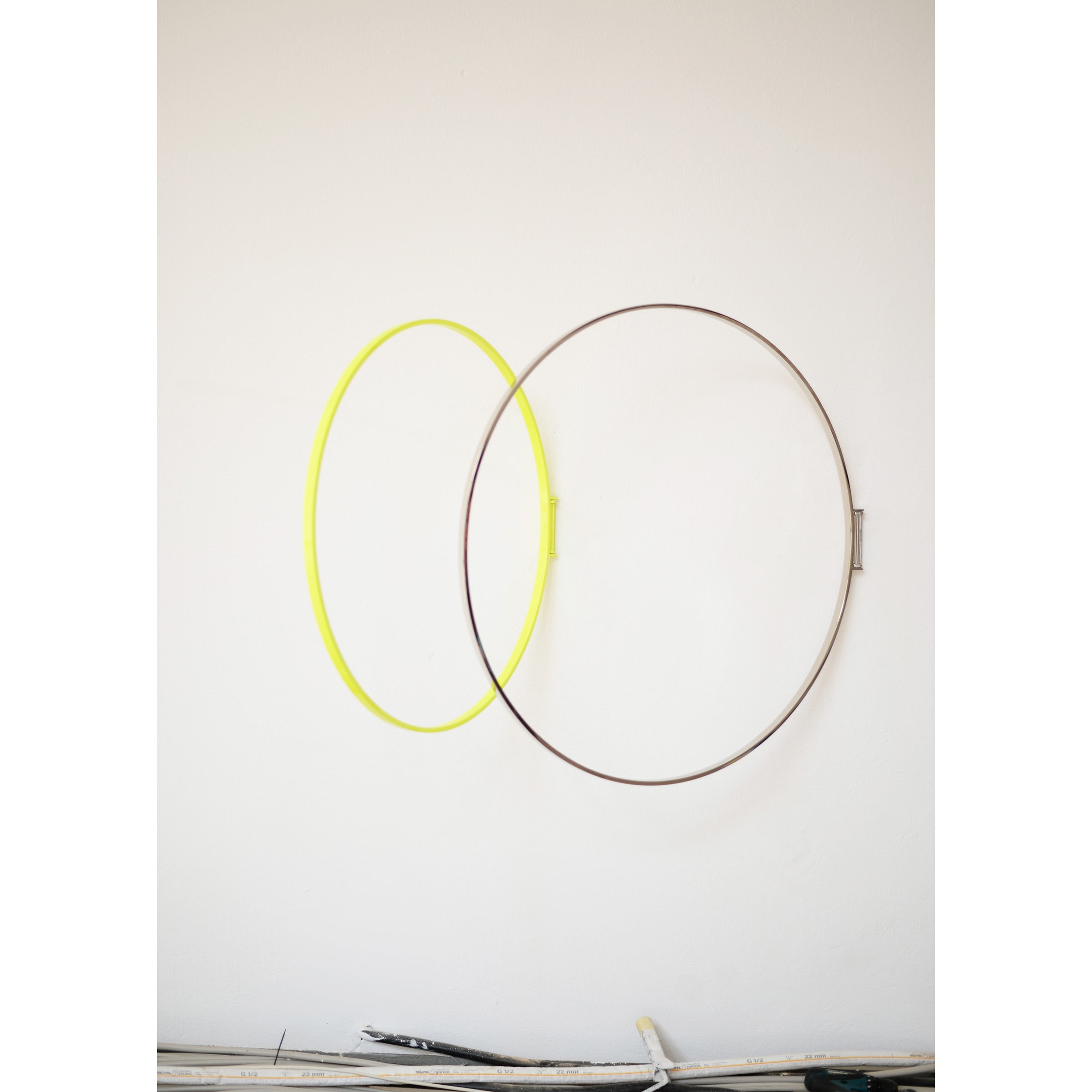 Atelier Haussmanns Rings, featuring intersecting yellow and brown metal rings, are mounted on a white wall. This decorative piece shows some visible scuffs, with a small ledge at the bottom holding assorted items.