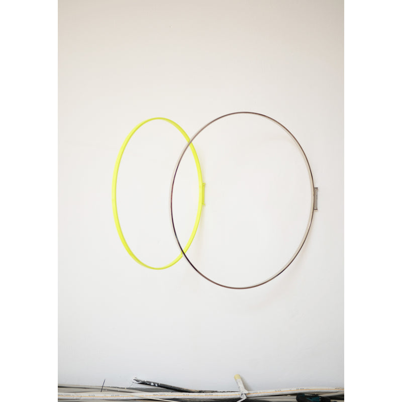Atelier Haussmanns Rings, featuring intersecting yellow and brown metal rings, are mounted on a white wall. This decorative piece shows some visible scuffs, with a small ledge at the bottom holding assorted items.
