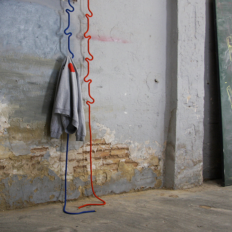 A gray sweatshirt hangs on a wavy, abstract Roberope coat rack with adjustable hooks, set against an aged wall with peeling paint and exposed bricks. The floor is concrete, and a large slate board stands nearby.