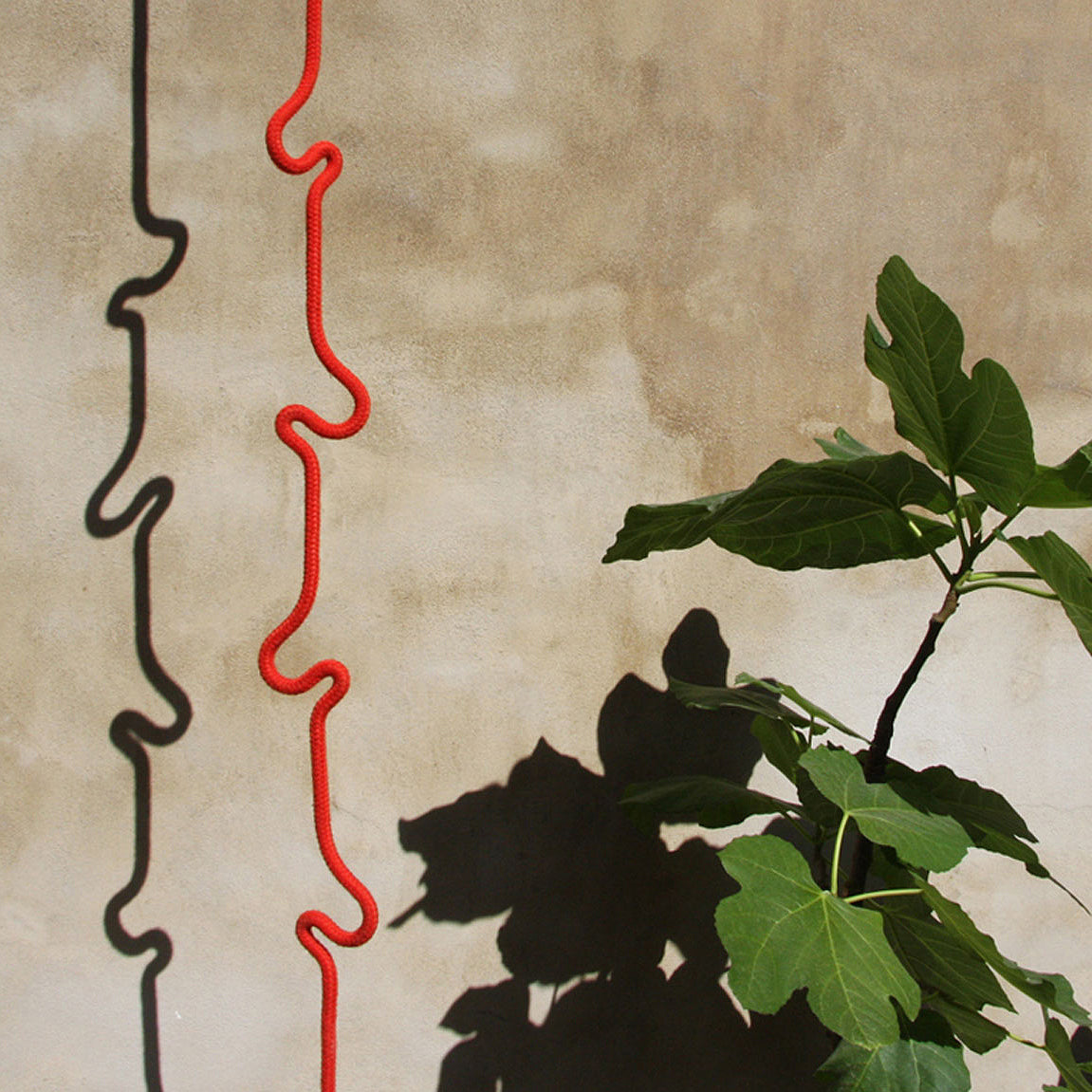 A red Roberope, crafted by the brand Roberope from polyester yarn, hangs against a beige concrete wall, casting a wiggly shadow. To its right, a green plant with large leaves projects its own artistic shadow on the wall.
