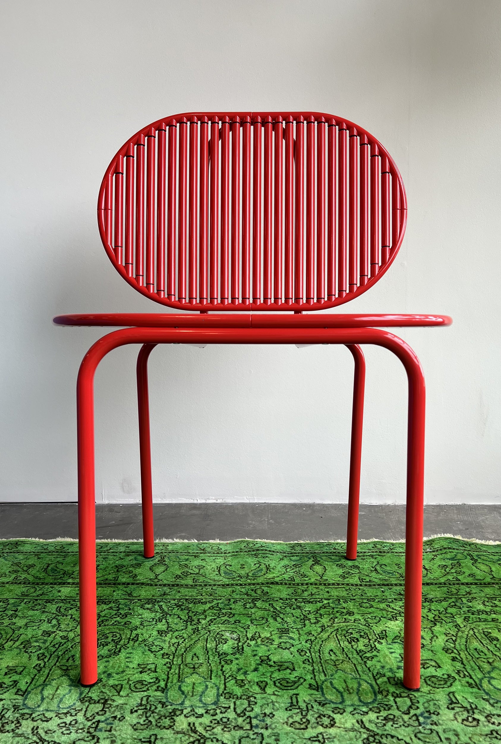 The AKTTEM Roll Chair - Sale, designed in Germany, boasts a modern red style with a circular slatted backrest and round seat. Its cylindrical legs of rotatable aluminum elegantly stand on a green patterned rug against a plain white wall.