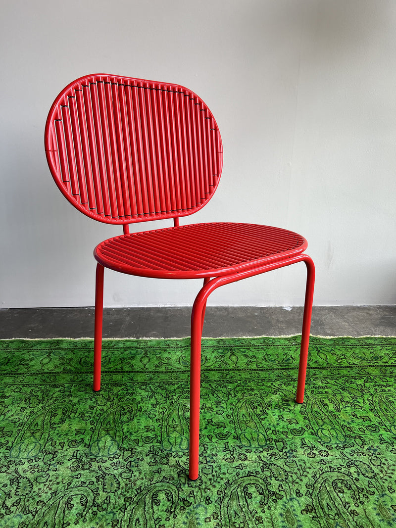 Introducing the Roll Chair - Sale by AKTTEM, a modern marvel with rotatable aluminum sticks in striking red. Designed in Germany, it boasts a round back and seat on sturdy tubular legs, displayed on a patterned green rug against a plain light-colored wall.