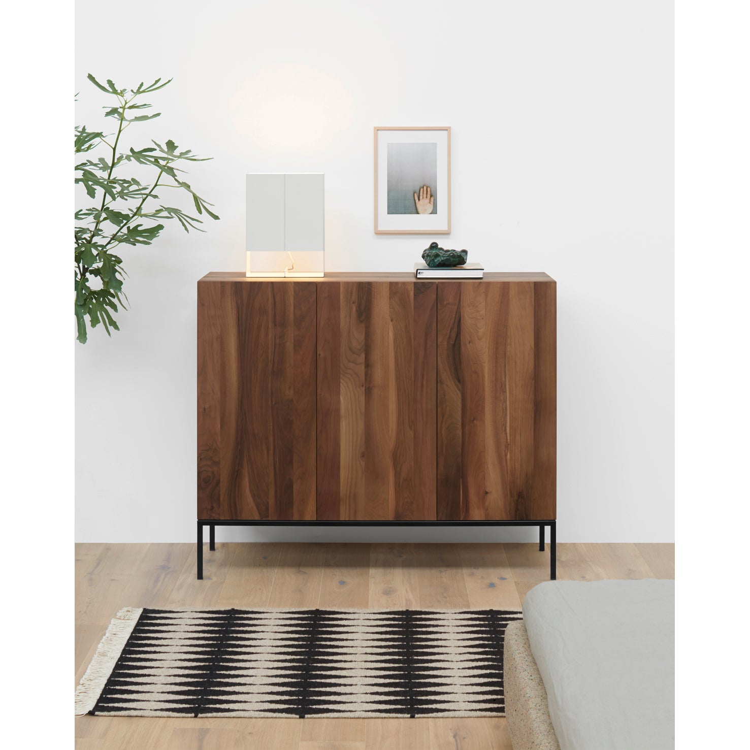 A minimalist room showcases the E15 SB03 FARIBA sideboard with black legs, topped by a leafy plant, lit lamp, and framed photo. A patterned rug enhances the wooden floor, with part of a bed visible in the lower right corner.