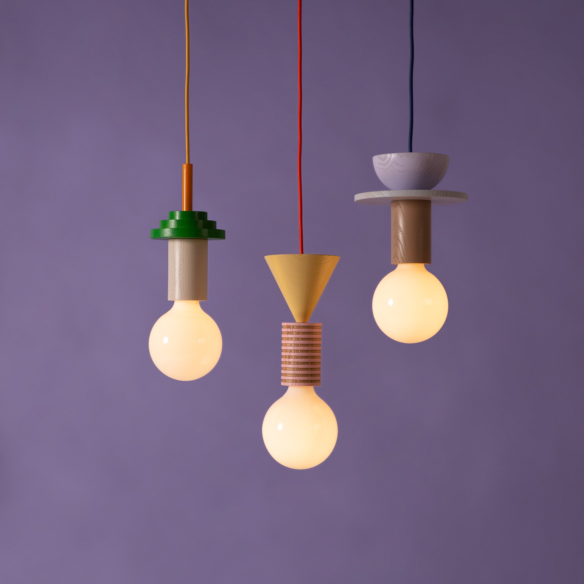 Three Junit Torta pendant lights by Schneid hang against a purple background, showcasing Bauhaus lighting. Each light features colorful geometric fixtures: a green disk, a yellow cone, and a purple dome, providing elegant ambient lighting.