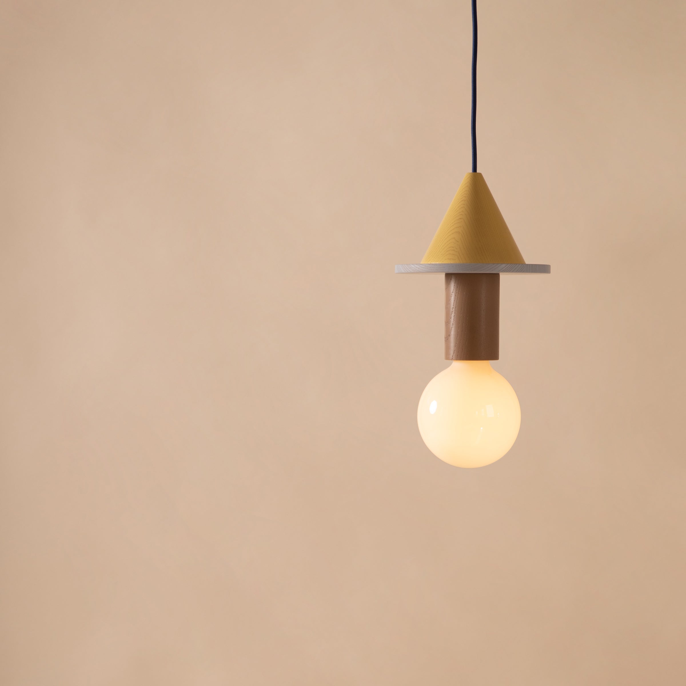 The Junit Meringe by Schneid is a Bauhaus-inspired pendant light featuring a yellow cone and round base with a glowing bulb on a black cord, set against a light brown background. Its minimalist design and soft ambient light make it ideal for contemporary spaces.