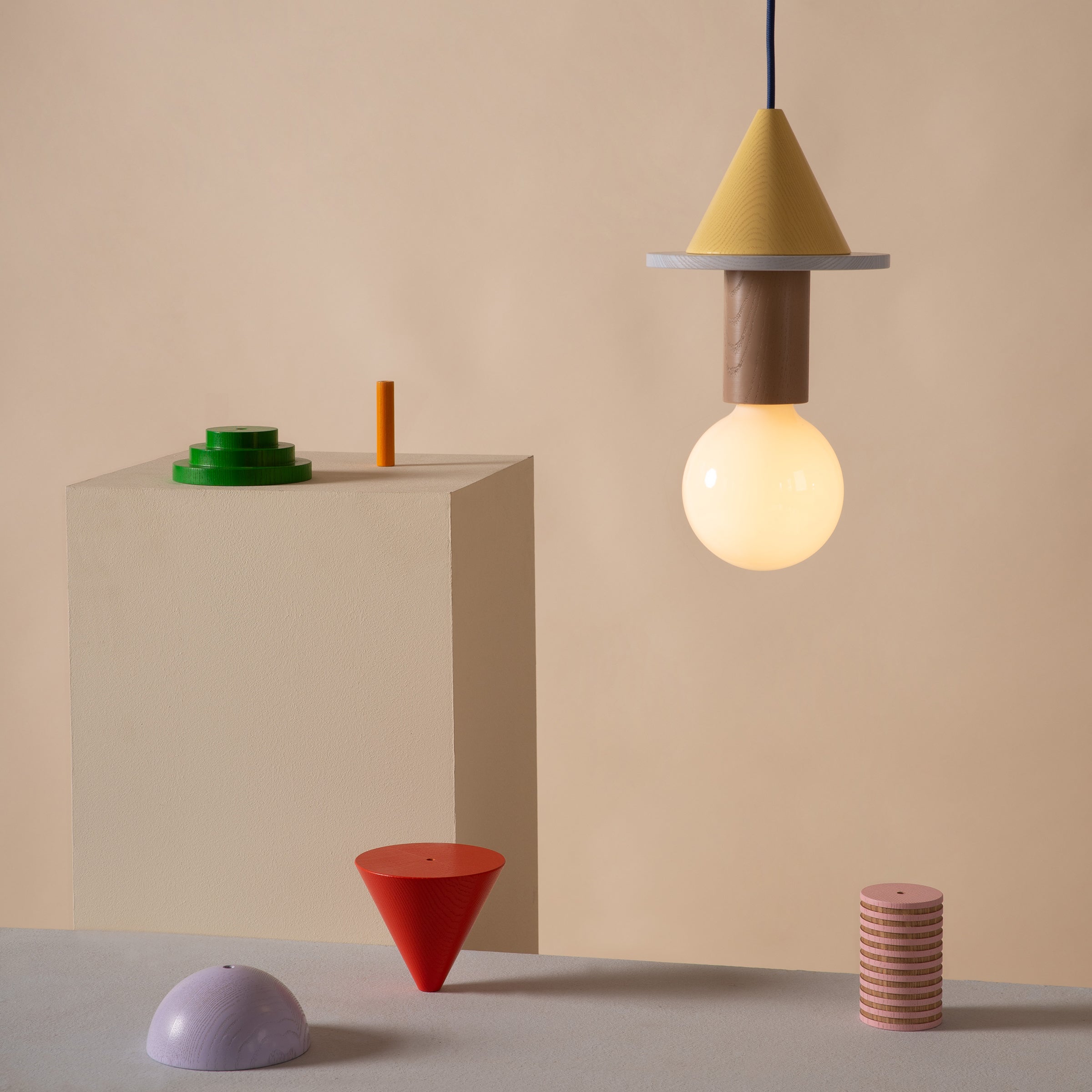 In a minimalistic, Bauhaus-inspired scene, Schneids Junit Meringe lamp hangs centrally, featuring ambient light with a glowing bulb and circular shade beneath its conical top. Below, vibrant geometric forms—a red cone, purple half-sphere, and striped cylinder—are elegantly arranged on the surface.