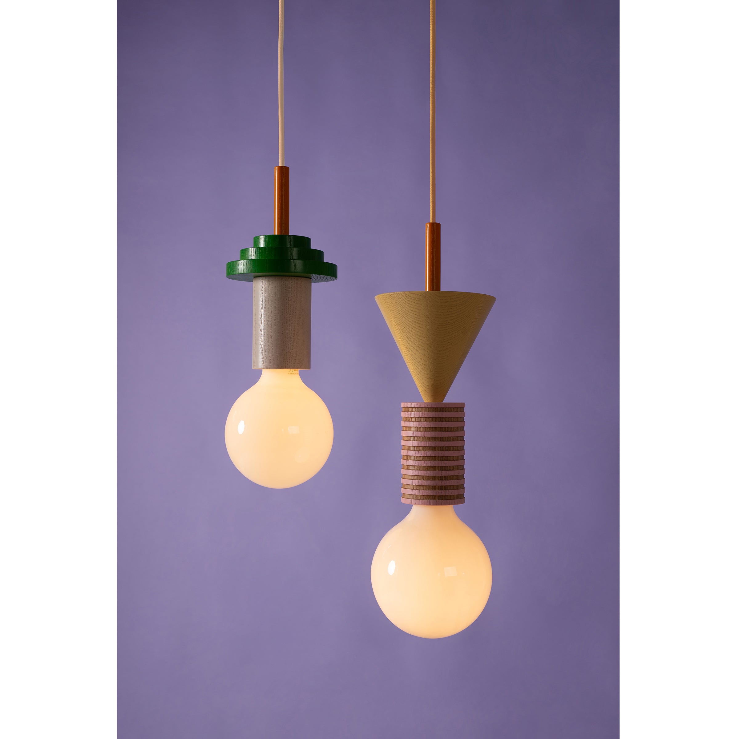 Two modern pendant lights, the Junit Fruta by Schneid, hang against a purple backdrop with Bauhaus influences. The left has a green top and cylindrical base, while the right features a conical top with ribbed geometric shapes. Both emit a warm, ambient glow.