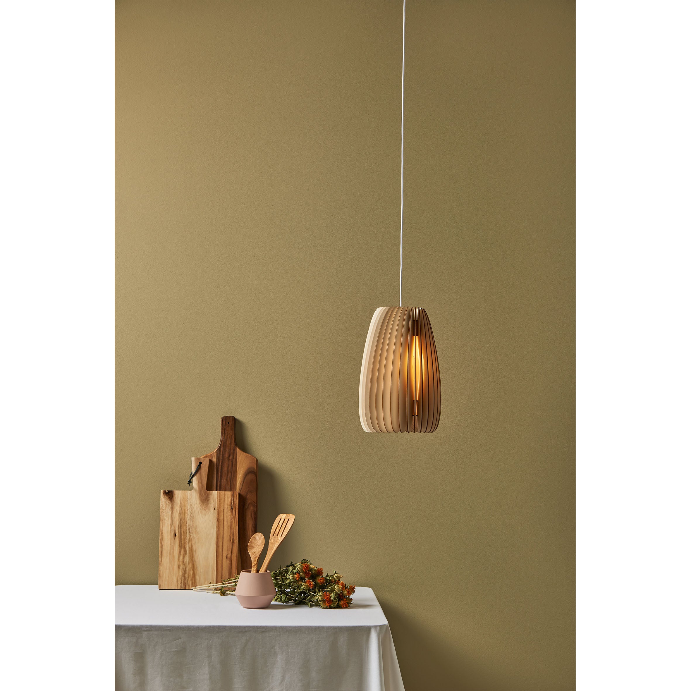 A modern Secundum pendant light by Schneid, made from poplar wood, illuminates an olive green wall. Below it sits a table with a white cloth, holding wooden cutting boards, a pink utensil container, and a small bouquet of orange and white flowers.
