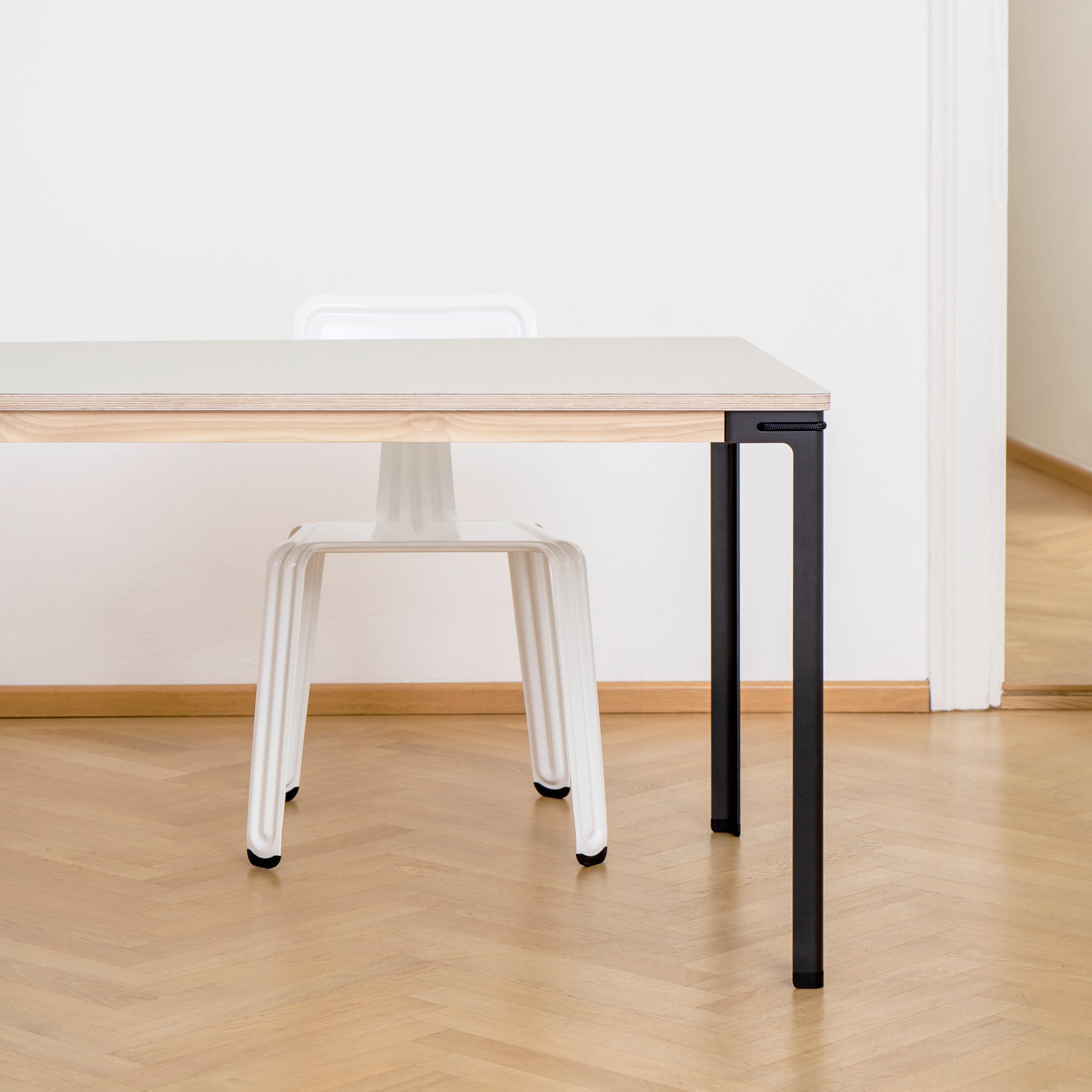 The minimalist room features a Moormann Seiltänzer Table with a white surface, light wood edge, and sleek steel legs. A white chair is partially tucked underneath on herringbone-patterned wooden flooring, set against a white wall with an open door.