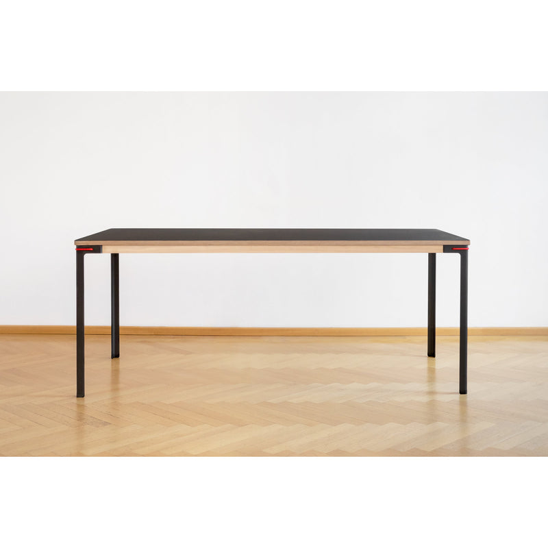 The Moormann Seiltänzer Table - SALE offers a minimalist design with a sleek black linoleum tabletop and thin steel legs, elegantly set against a white wall on a light wooden floor.