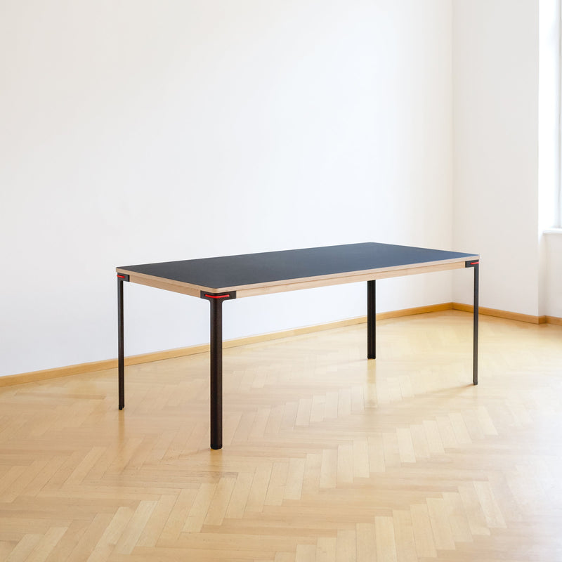 The Seiltänzer Table by Moormann, currently on sale, showcases a dark linoleum rectangular top with light wood edges and slender steel legs. It is elegantly set on polished herringbone flooring, in a bright room with white walls and a right-side window.