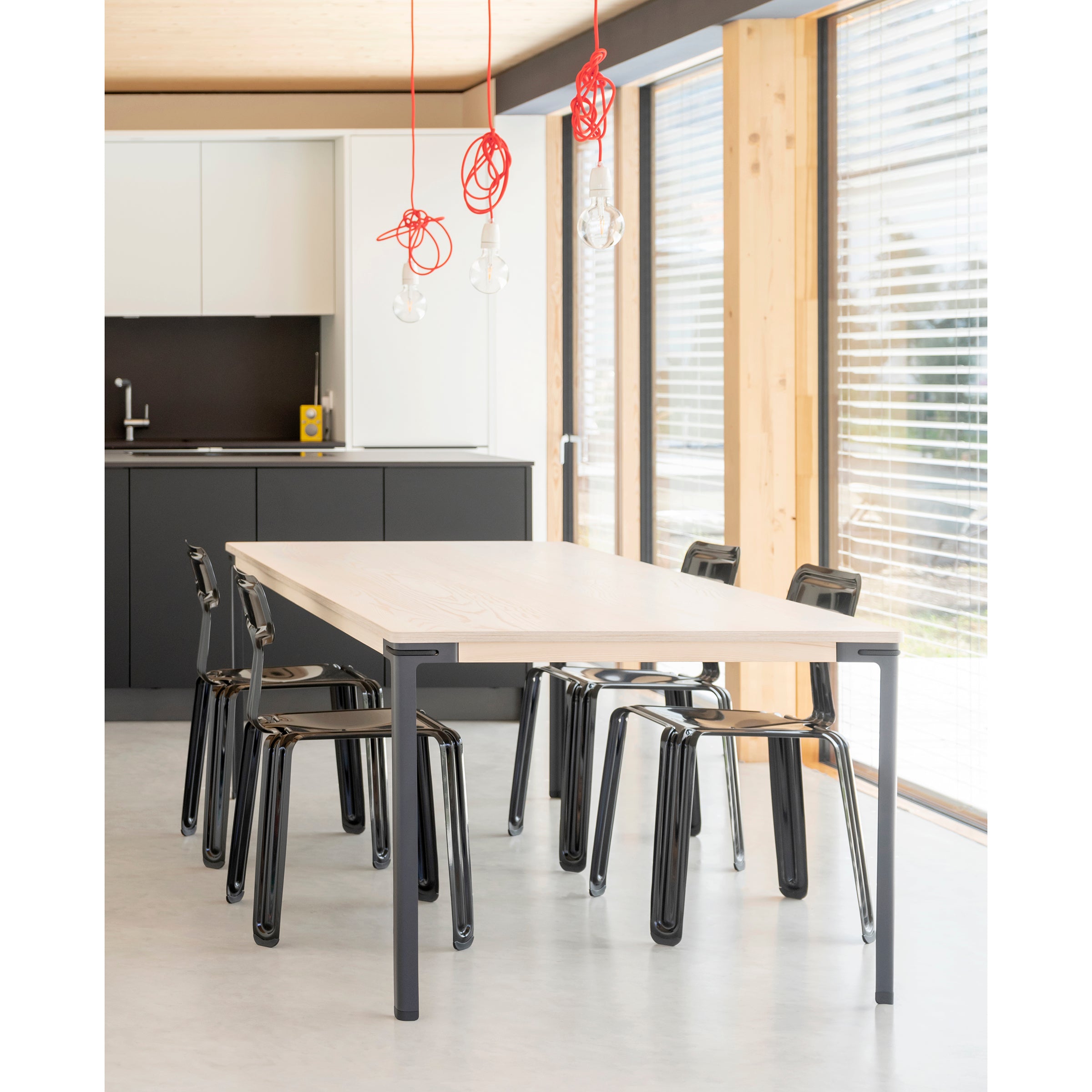 A modern dining area features a Moormann Seiltänzer Table with sleek steel legs and six black chairs, illuminated by hanging red cords with light bulbs. The backdrop includes a minimalist kitchen and large windows with blinds, allowing natural light to enhance the space.