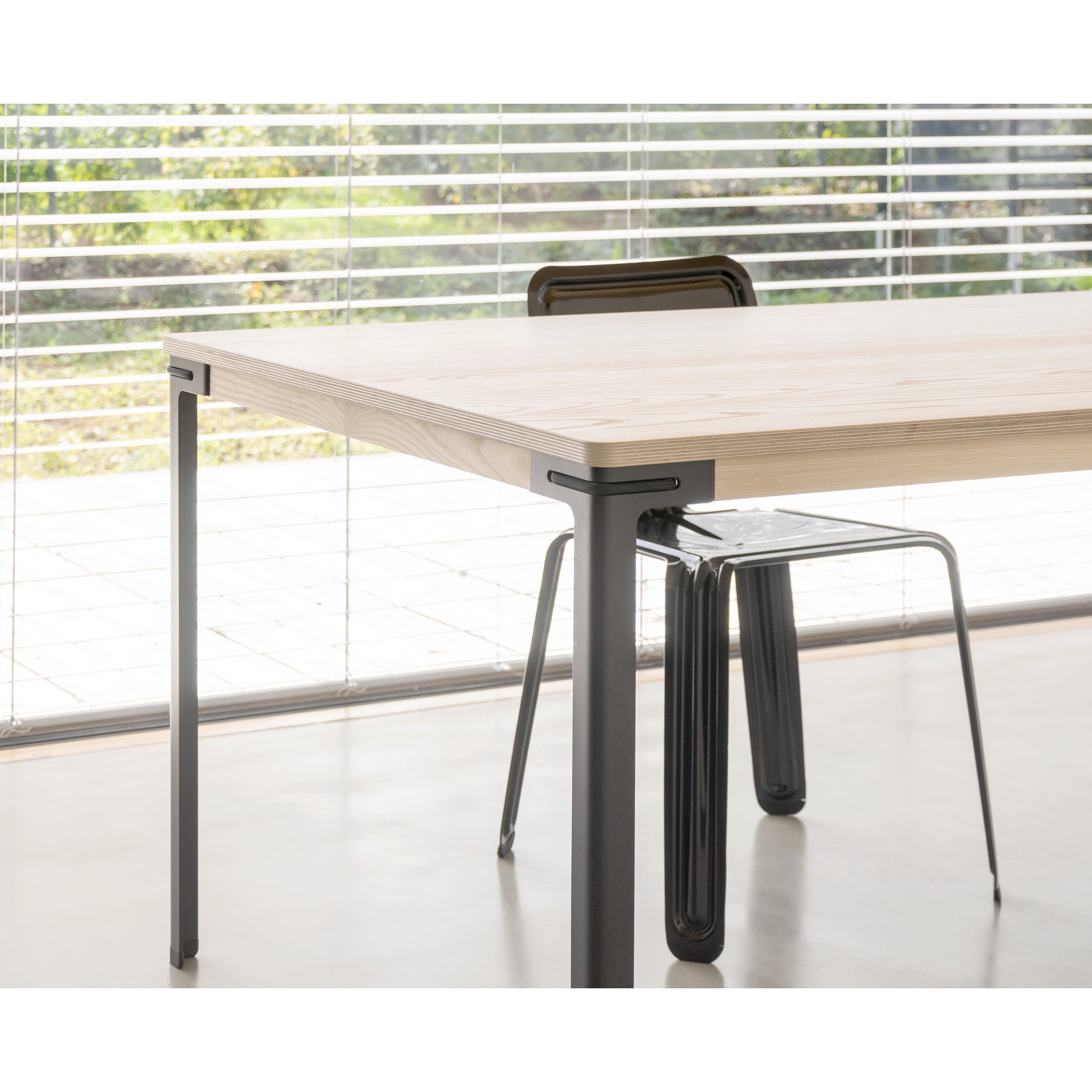 A modern dining area features a Seiltänzer Table by Moormann with sleek steel legs. Large windows with horizontal blinds reveal a sunny garden view, while the minimalist space boasts clean lines and a neutral color palette enhanced by its modular design.