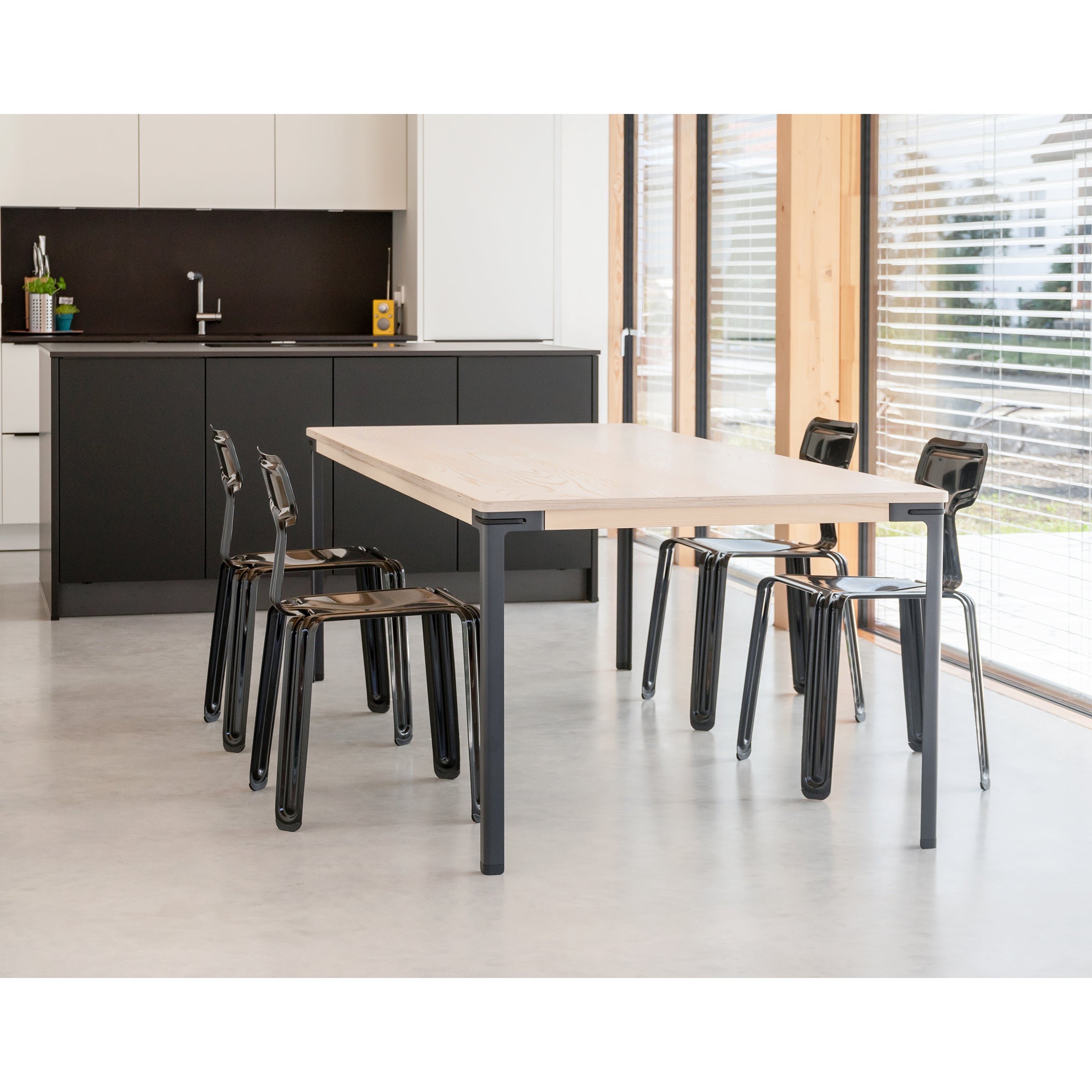 A modern kitchen with a sleek, modular design features the Seiltänzer Table by Moormann, boasting wooden surfaces with steel legs and black metal chairs. Black cabinets complement the sink and countertop accessories, while large windows with blinds invite natural light into the space.