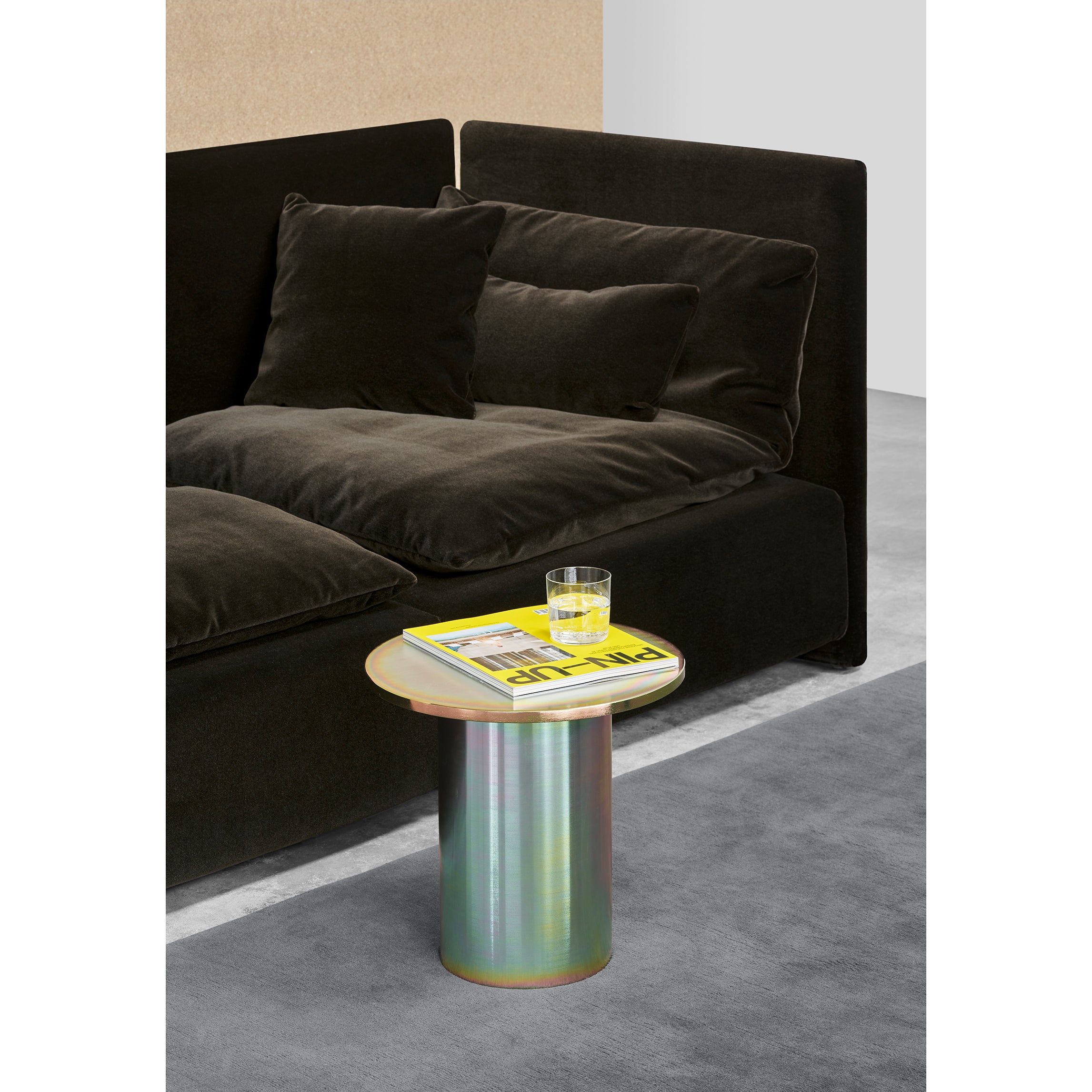 A modern, plush black sofa with several cushions sits next to a sleek CT09 ENOKI METAL side table by E15, featuring a powder-coated steel finish. The table holds a glass of yellow drink and a magazine on the gray carpeted floor in the minimalist room.