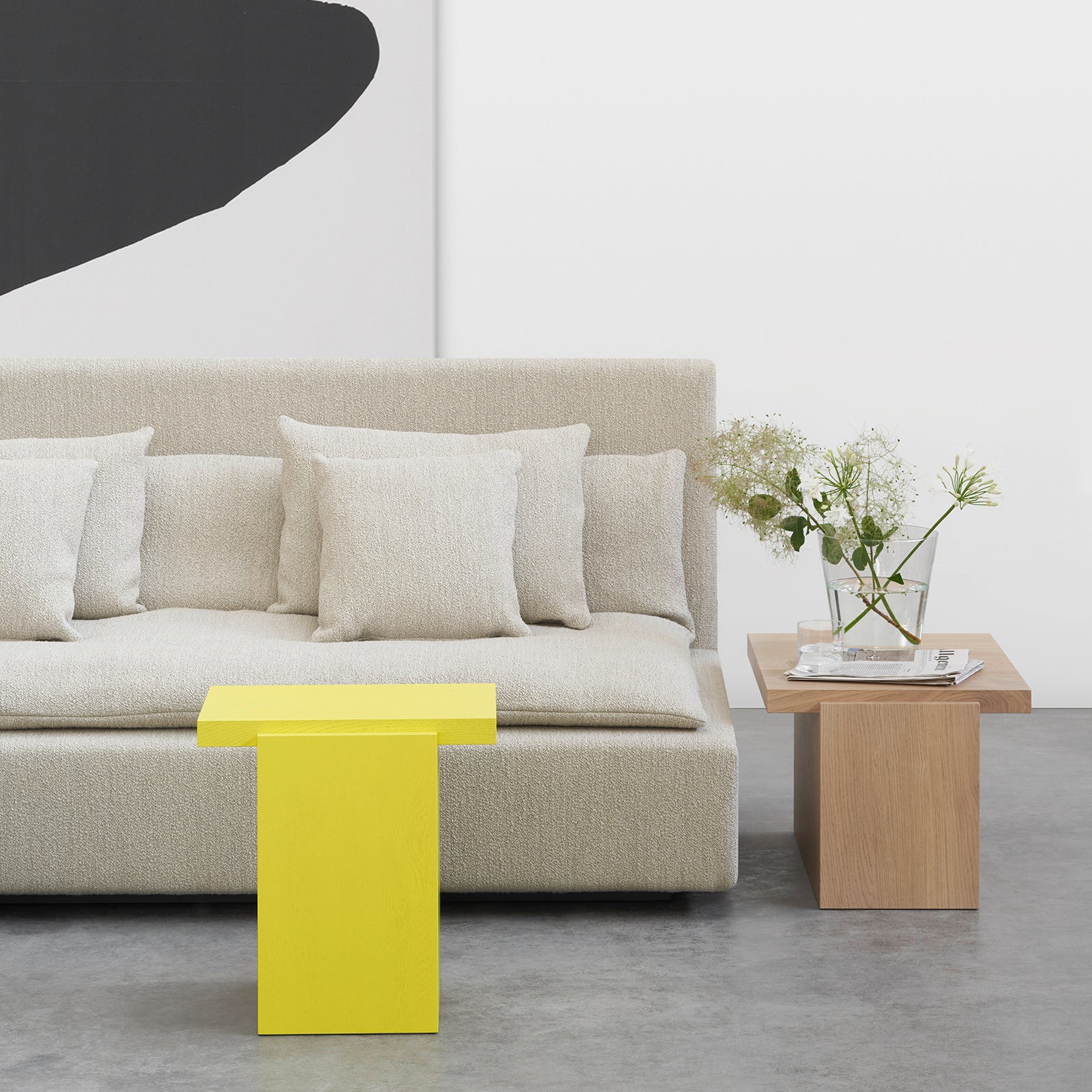 A minimalist living room features a light beige sofa with cushions. A bright yellow coffee table and E15s solid wood DT02 TORE side tables hold a vase of white flowers. A large black and white abstract artwork decorates the wall.