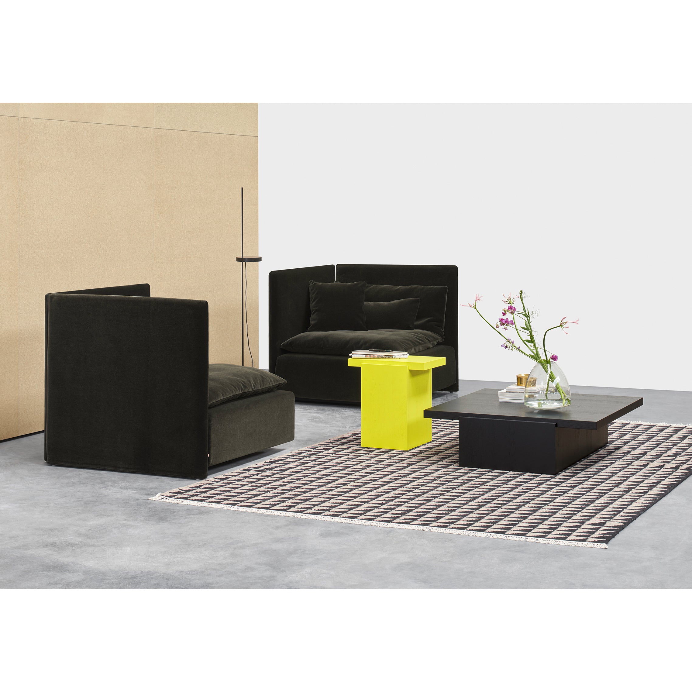 A modern living room features two black armchairs, a bright yellow side table, and a low black coffee table on a patterned rug. A minimalist floor lamp and vase with flowers adorn the DT02 TORE marble side tables by E15. Neutral walls and a gray floor complete the setting.