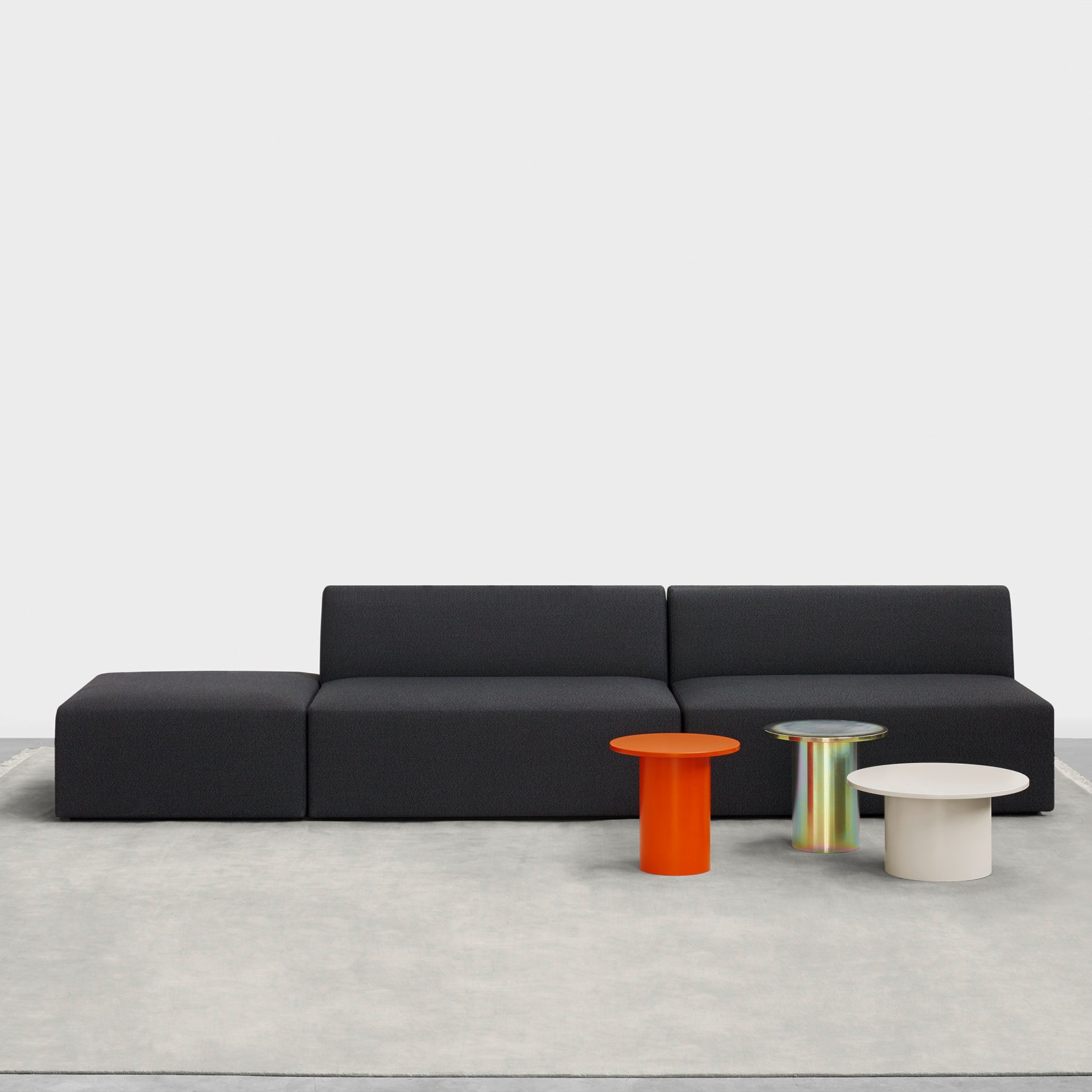 A sleek, modern living room setup showcases a dark gray modular sofa on a light gray rug. In front are three round tables: one orange, one iridescent, and the CT09 ENOKI METAL side table in cream by E15 with powder-coated steel details by Philipp Mainzer, adding pop to the minimalist design.