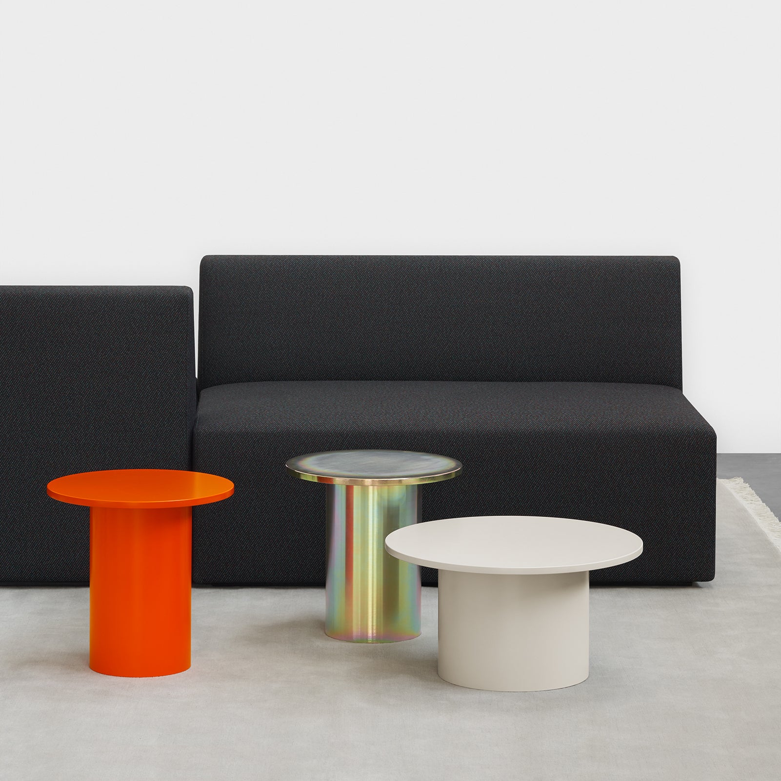A modern interior highlights a minimalist black sofa against a white wall. In front, three elegant tables, including the iridescent CT09 ENOKI METAL side table by E15, rest on a light gray rug. The powder-coated steel design enhances the sleek sophistication of the space.