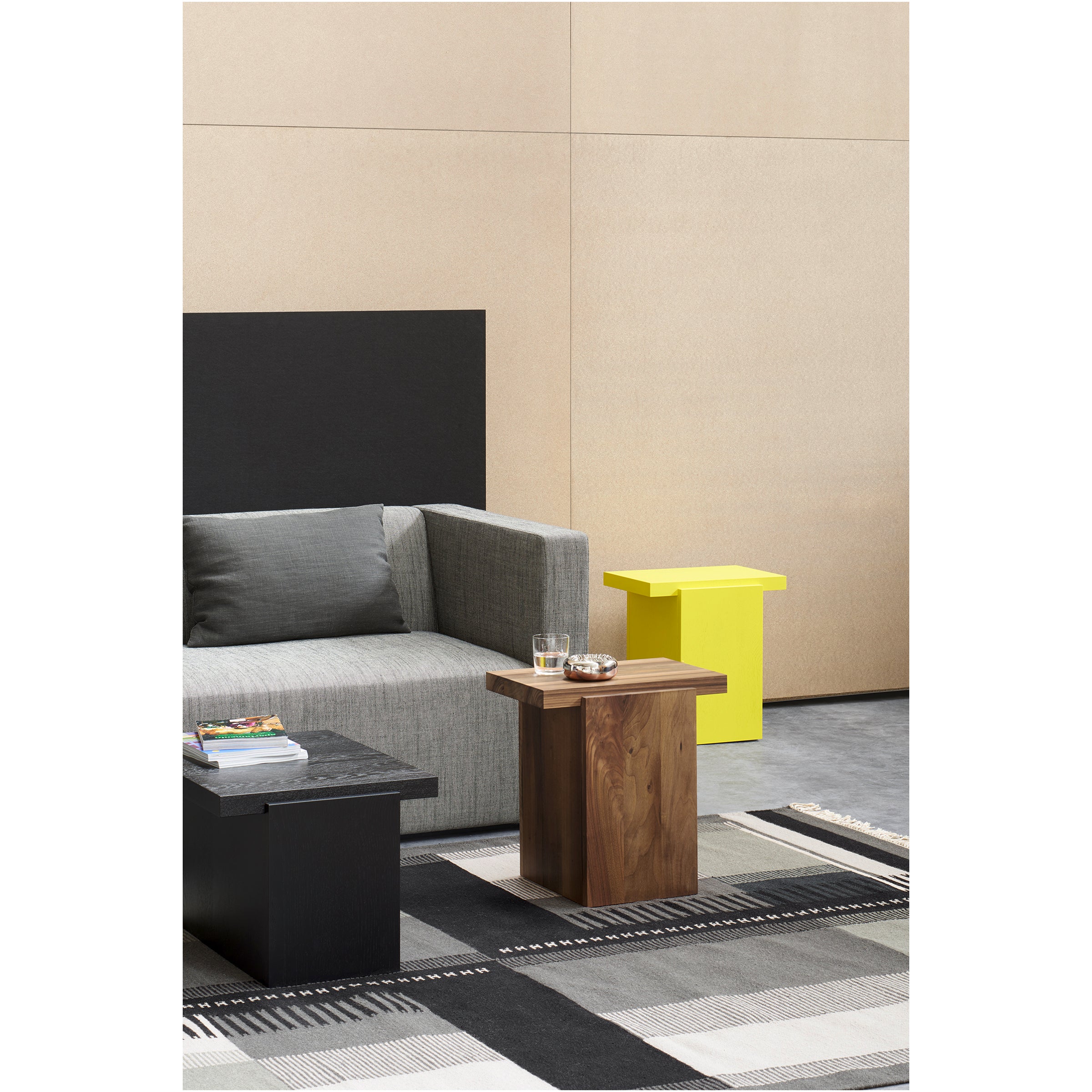 A modern living room showcases a gray sofa with sculptural side tables, including the E15 DT02 TORE. A yellow accent table adds color, and a checkered rug covers the floor. Books, a glass, and a small bowl sit on one TORE side table. The walls feature an elegant beige and black finish.