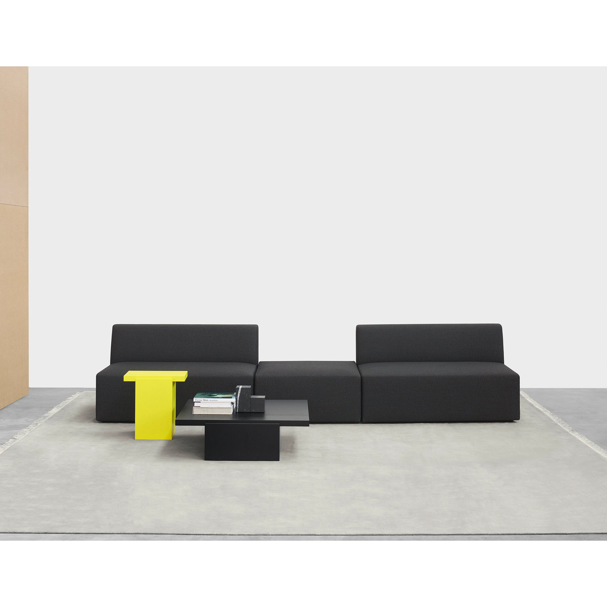 Three modern black sofas are arranged in a minimalist setting on a light gray rug, complemented by a bright yellow sculptural side table and a black coffee table with books and a small object. The contemporary design is accentuated by the elegant presence of E15s DT02 TORE side tables.