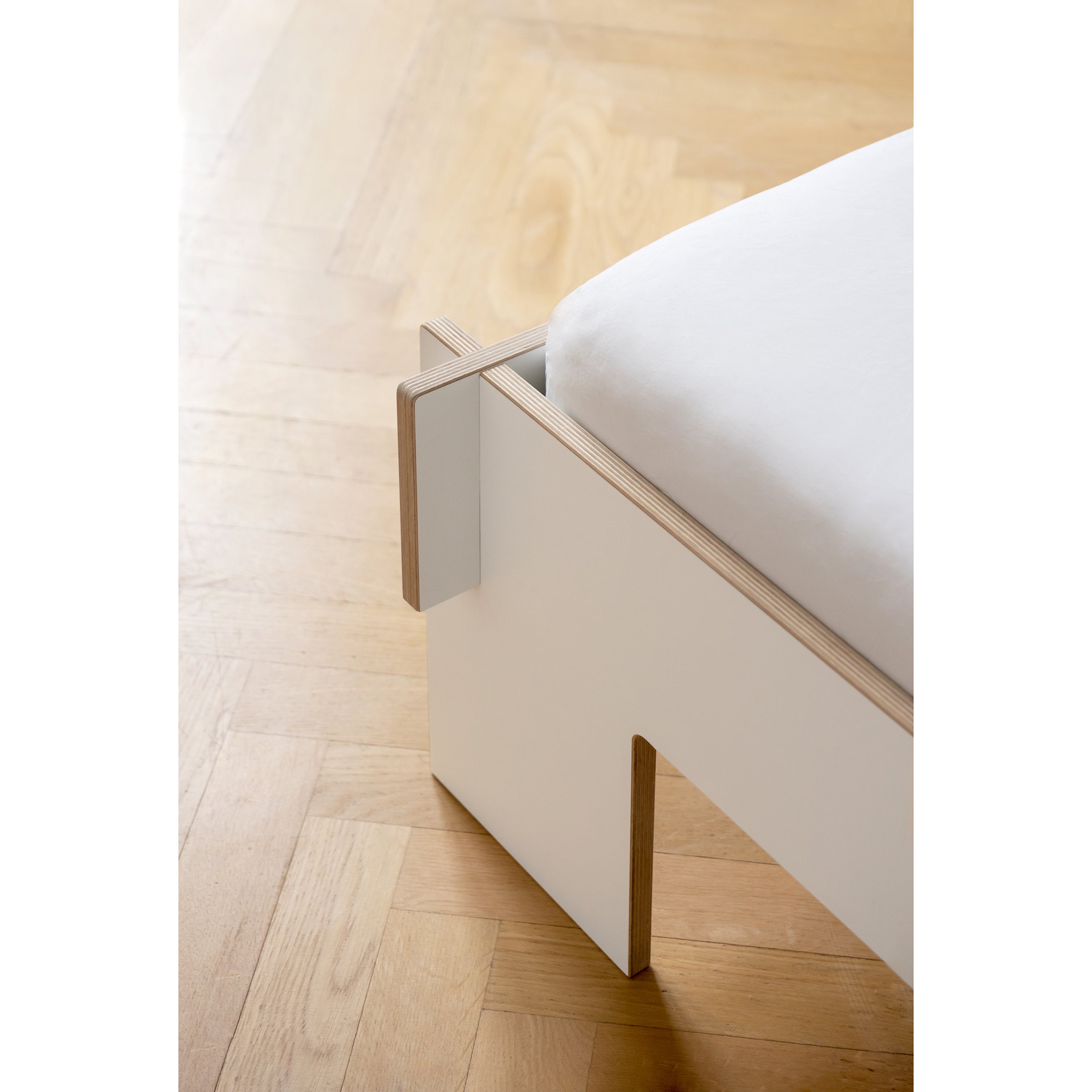 Close-up of the Siebenschläfer by Moormann, a minimalist white corner bed frame made from birch plywood with light wood edges on polished wooden floors. The flat-packed design features a clean, modern look with a fitted white sheet, and natural light highlights the wood grain and floor texture.