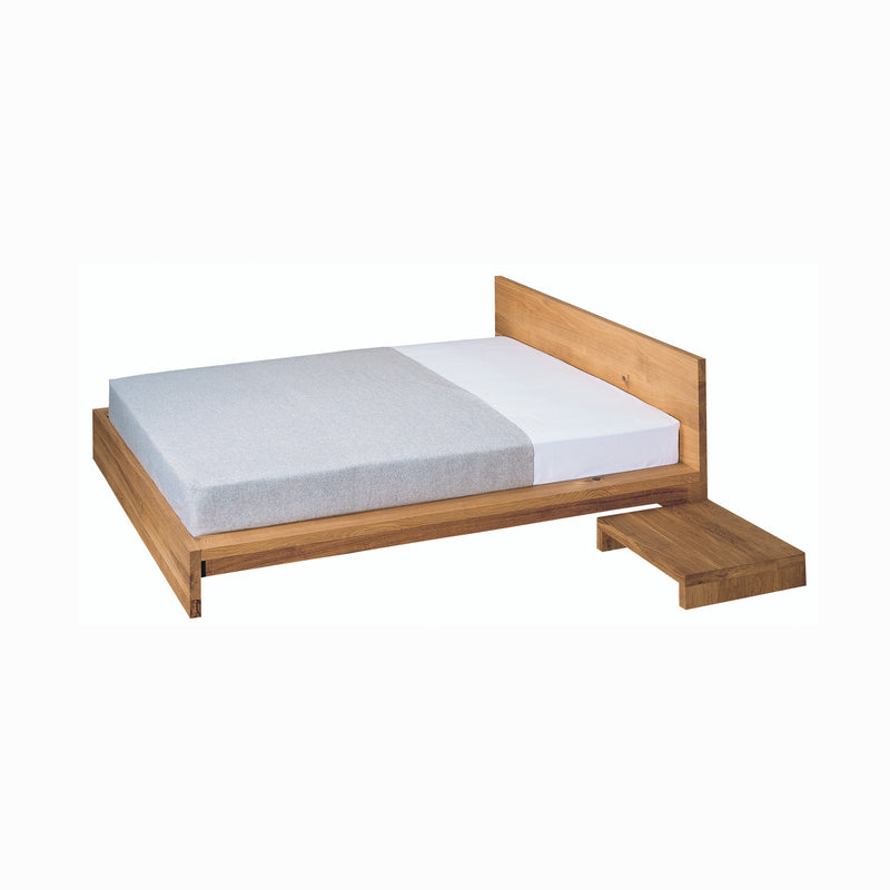 The NT01 MAX by E15 is a minimalist wooden bed crafted from solid wood, paired with a light gray mattress and a white pillow. A low bedside table complements the beds modern, sleek design.