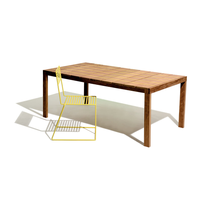 The Egoe Sena Table, crafted from tropical wood with a grid pattern, is paired with a yellow metal chair on one side, casting shadows on a light background for a timeless aesthetic.