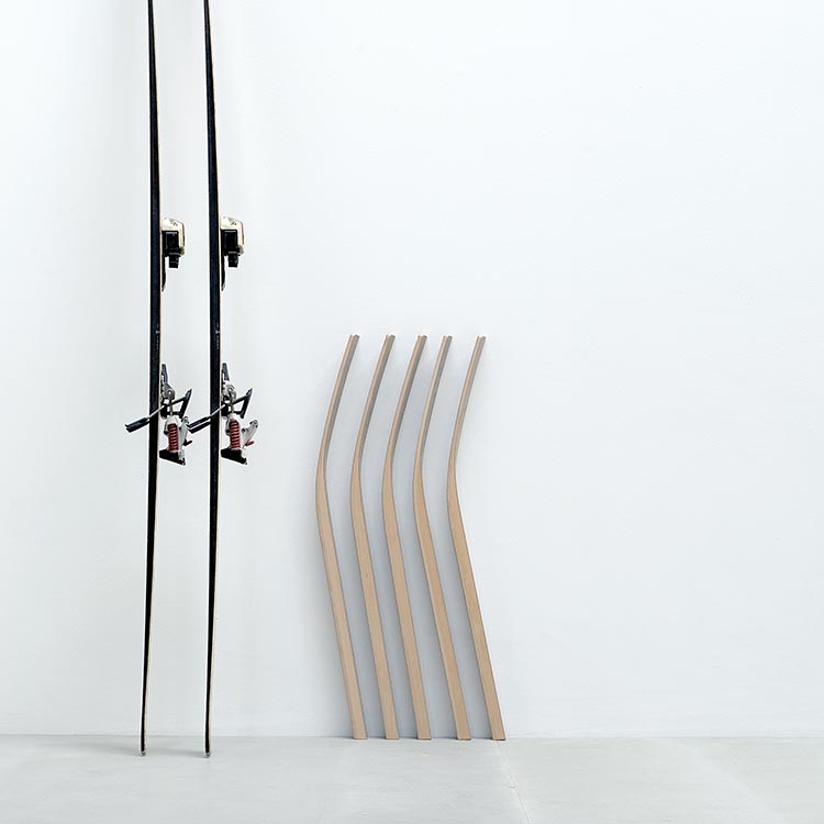 Two Hussl ST10 black skis lean against a white wall on the left, with five wooden composite curved planks beside them. This minimalist scene features a plain white background.