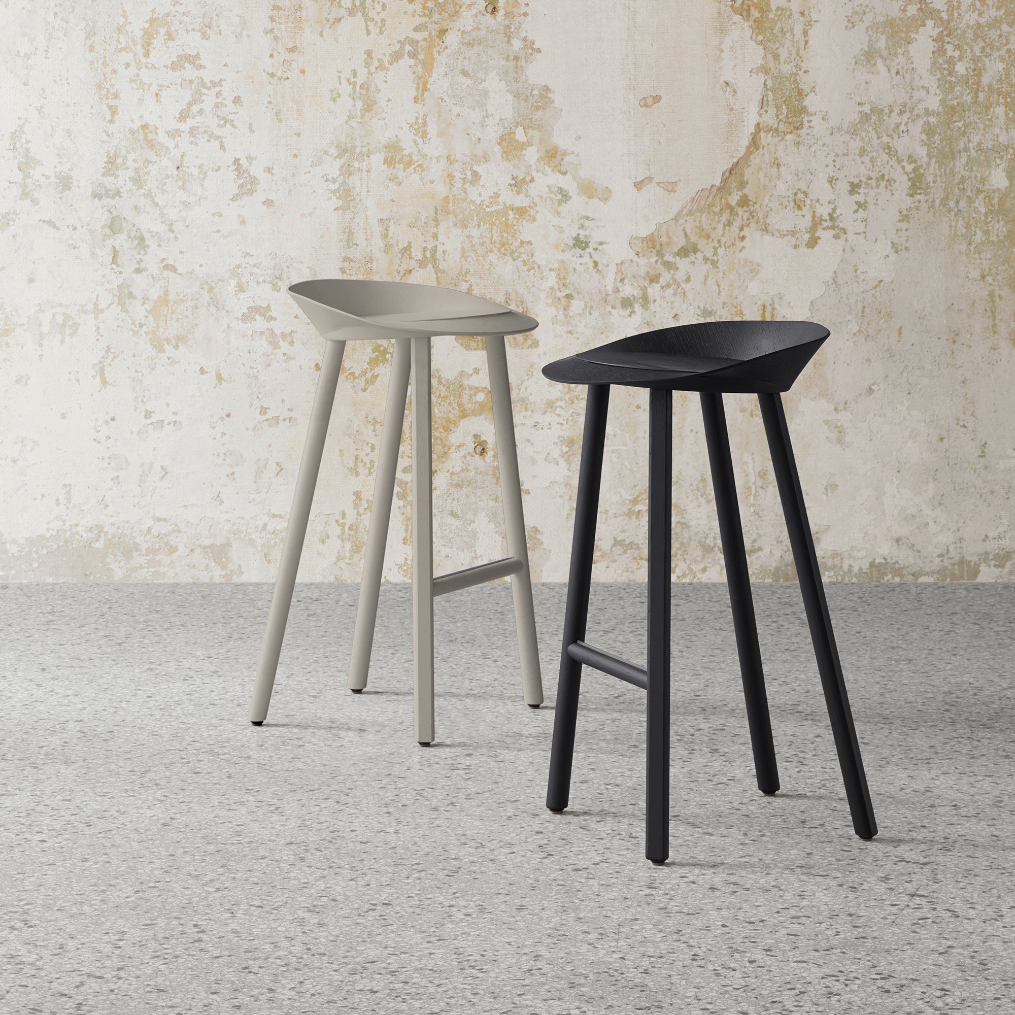 The ST10 JEAN bar stools by E15, with triangular seats made of oak-veneered plywood, feature three legs. One is light gray, the other black, set on a speckled gray floor against a textured wall with peeling beige and white paint.