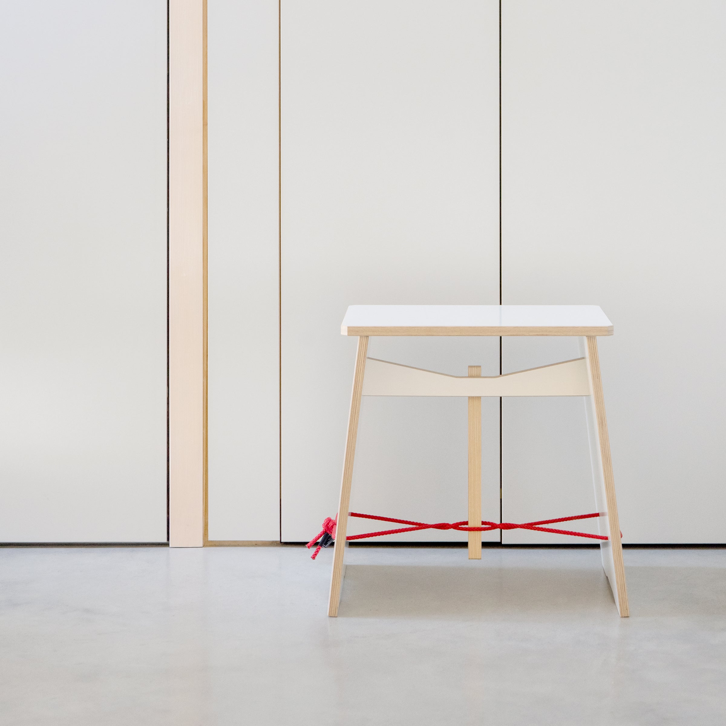 The minimalist Strammer Max table by Moormann, featuring a distinctive red cross-brace and a light wooden frame, stands on light gray flooring. In the background, white plywood panels with thin wooden accents enhance the rooms creative ambiance.