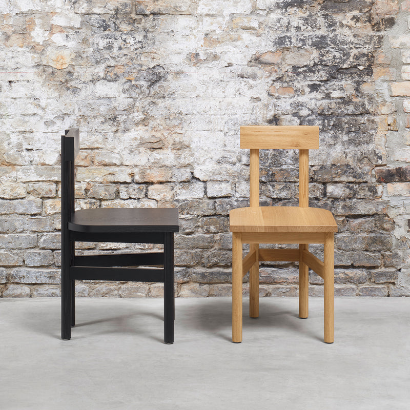 The GAMAR collection by E15 features the SX01 GAMAR Chair in two finishes: black and natural European oak. These rustic wooden chairs, resting on a concrete floor against a textured brick wall, showcase a minimalist design with straight lines and square backs.