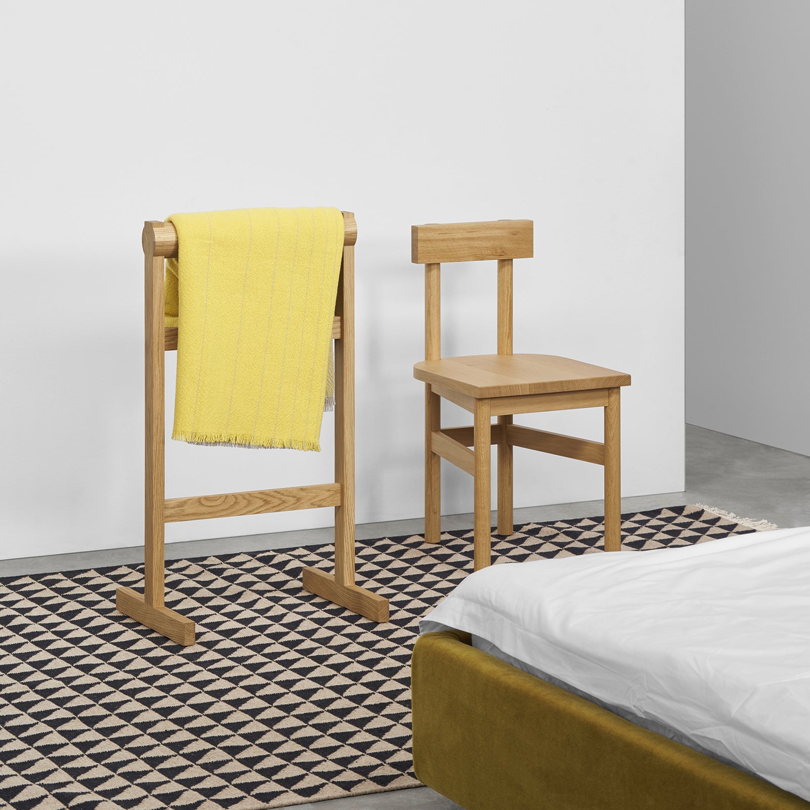 A minimalist room features an E15 SX01 GAMAR Chair and valet stand. A bright yellow towel hangs on the valet, which stands on a geometric rug. A bed with white bedding is partially visible against a plain white wall.