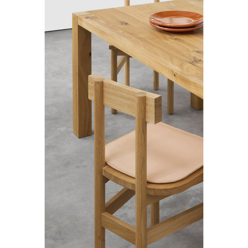 An SX01 GAMAR rustic wooden chair with a light tan seat from E15 is placed next to a wooden dining table. Two ceramic orange plates from the GAMAR collection sit on the table. The gray floor has a smooth texture.