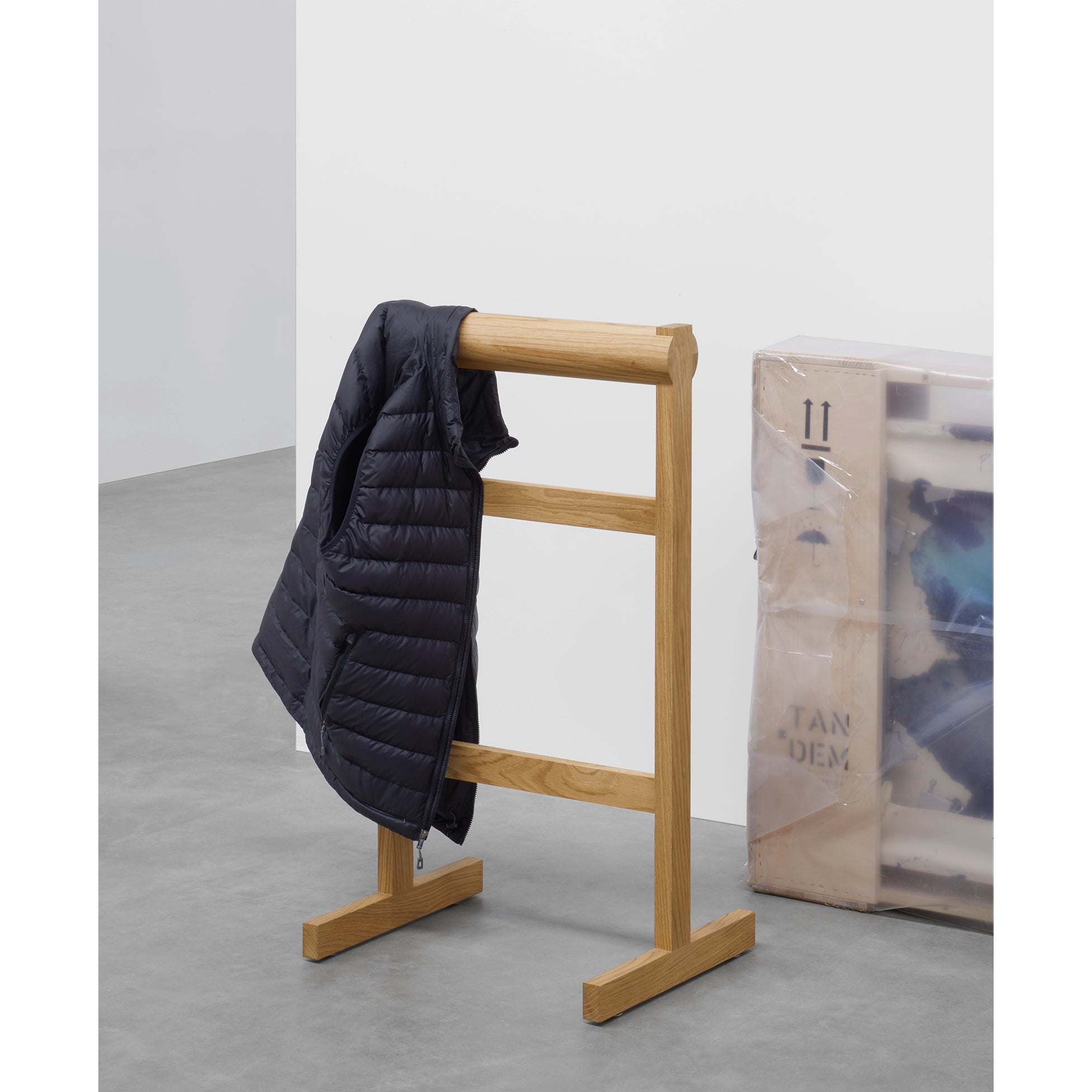 The SX03 GAMAR Wardrobe Stand by E15, made of European oak, holds a black puffer jacket on a gray concrete floor. A leaning wrapped canvas completes the rooms understated elegance.