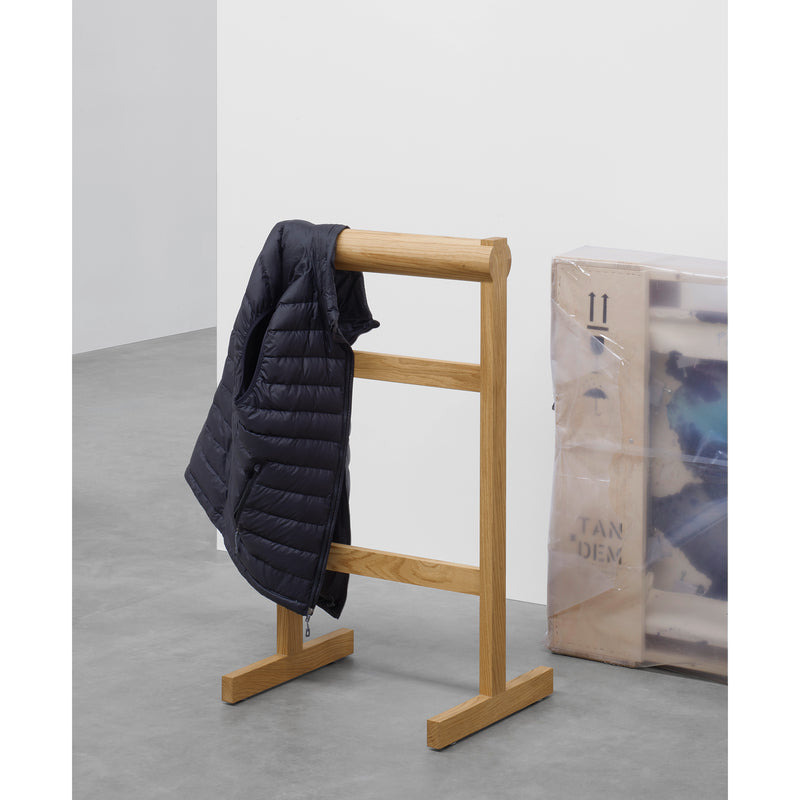 The SX03 GAMAR Wardrobe Stand by E15, made of European oak, holds a black puffer jacket on a gray concrete floor. A leaning wrapped canvas completes the rooms understated elegance.