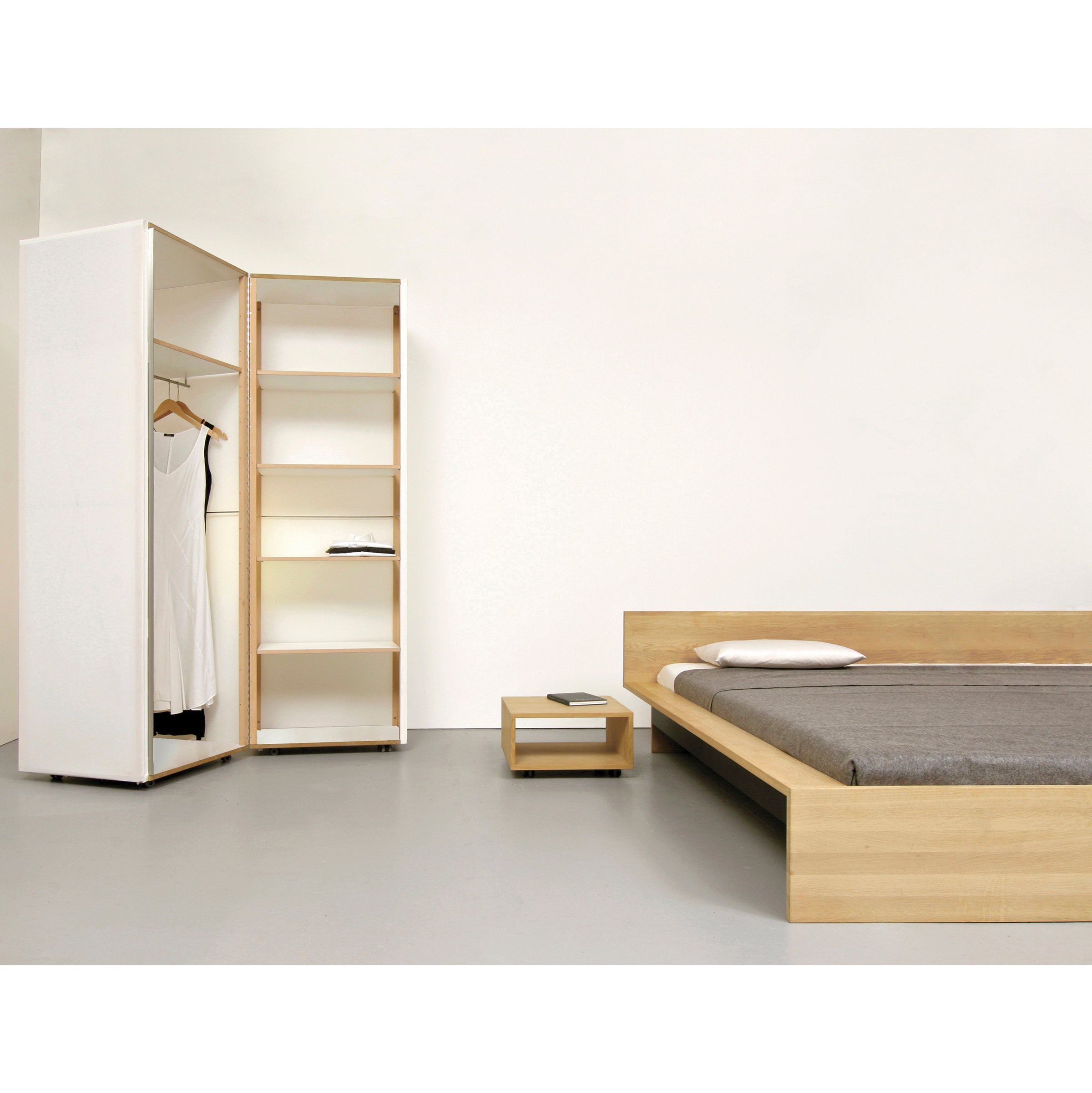A minimalist bedroom showcases the Sanktjohanser Dinadrei Bed with a high headboard, light wood, and gray bedding. A wooden nightstand holds a book, while an open wardrobe displays shelves and a white dress. The room features white walls and a sleek, modern design.