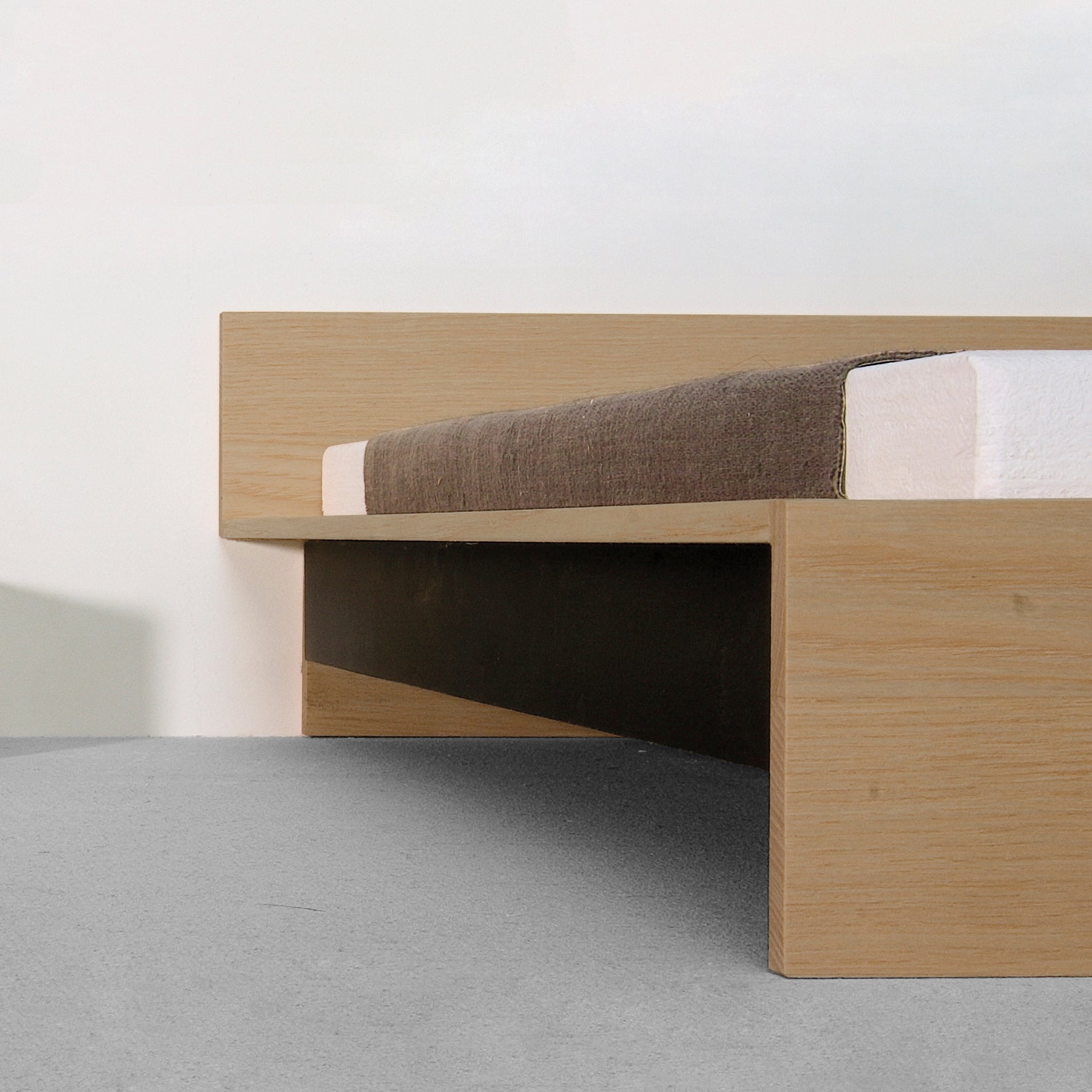 The Dinadrei Bed by Sanktjohanser is a minimalist solid wood platform bed with a simple, low rectangular headboard. The frame is light-colored wood, topped with a brown-covered mattress, set against neutral walls and floors.